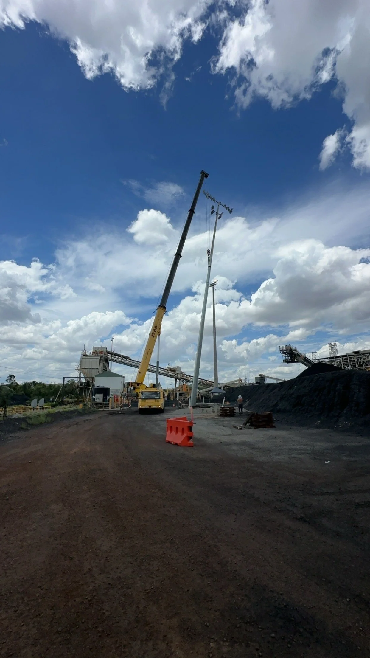 Construction site with a crane lifting a utility pole, construction materials, and workers under a partly cloudy sky.