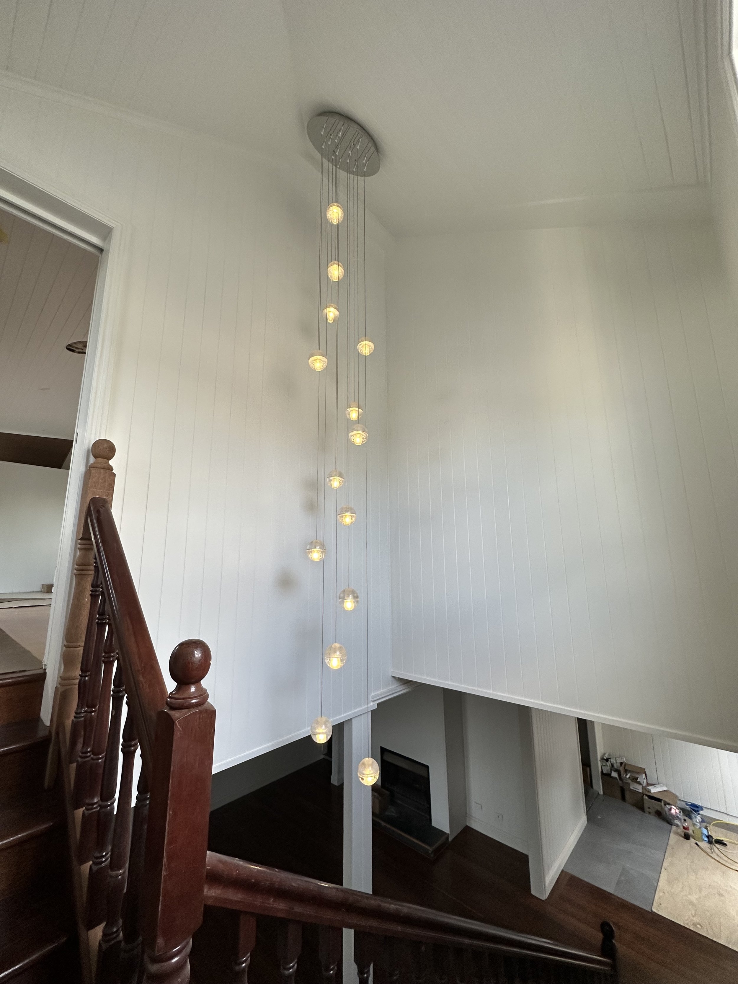 Interior view of a two-story staircase with a hanging modern chandelier, white vertical-paneled walls, and a corner with a kitchen or laundry area with tools and materials on the floor.
