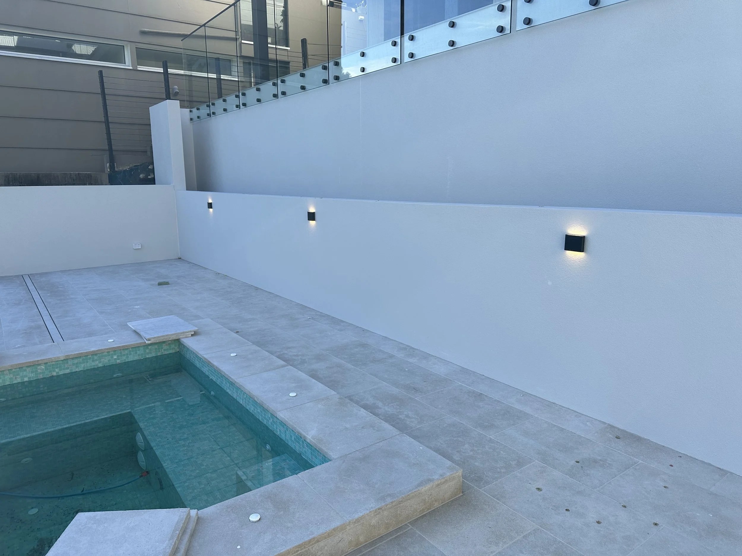 A modern outdoor patio with a small swimming pool, light gray tiled floor, white painted wall with modern black wall lights, and a glass railing on an upper level of a multi-story building.