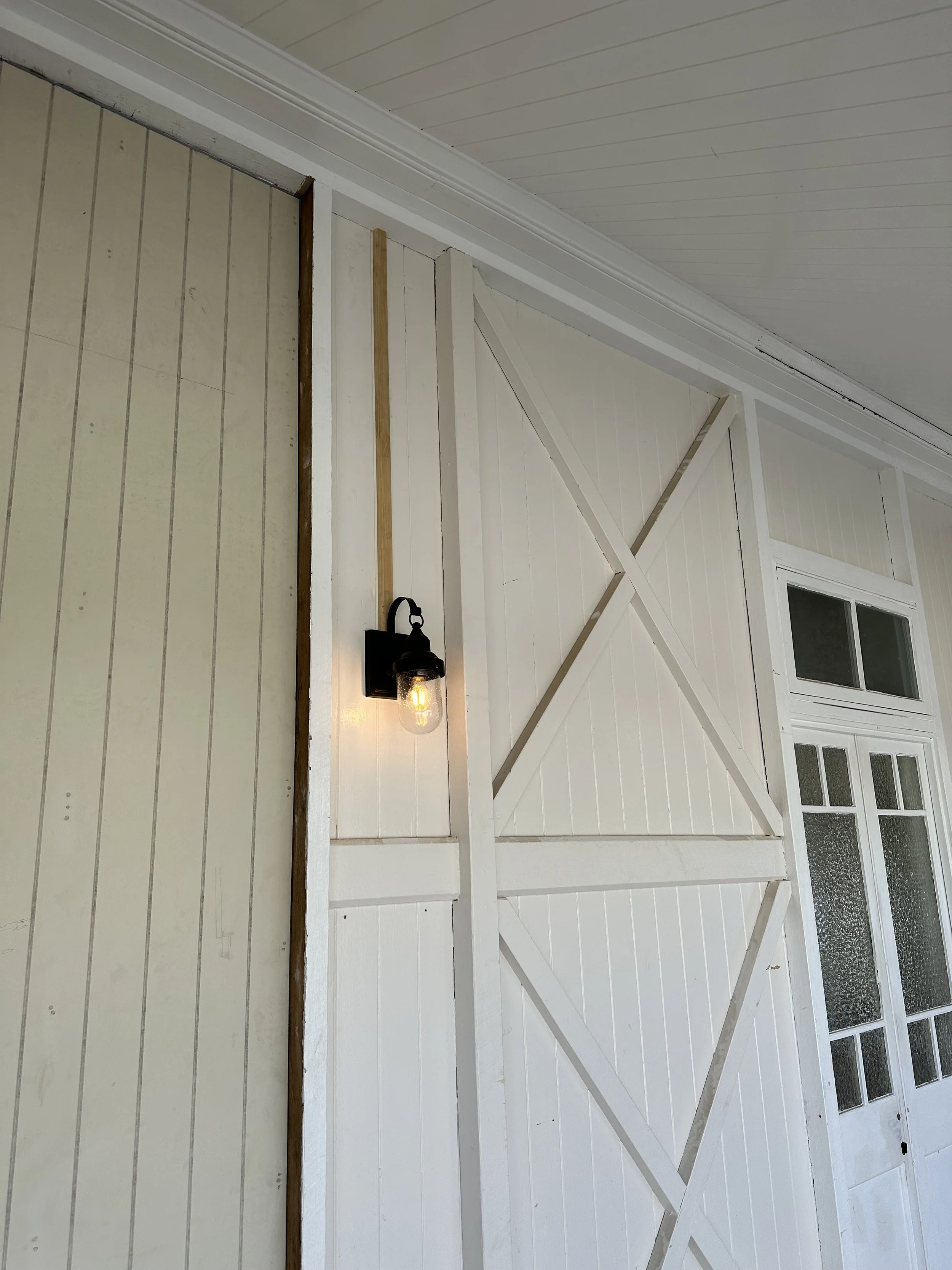 White wooden wall with a window, a black outdoor lantern light, and wooden cross braces. Part of a ceiling with horizontal panels is visible.