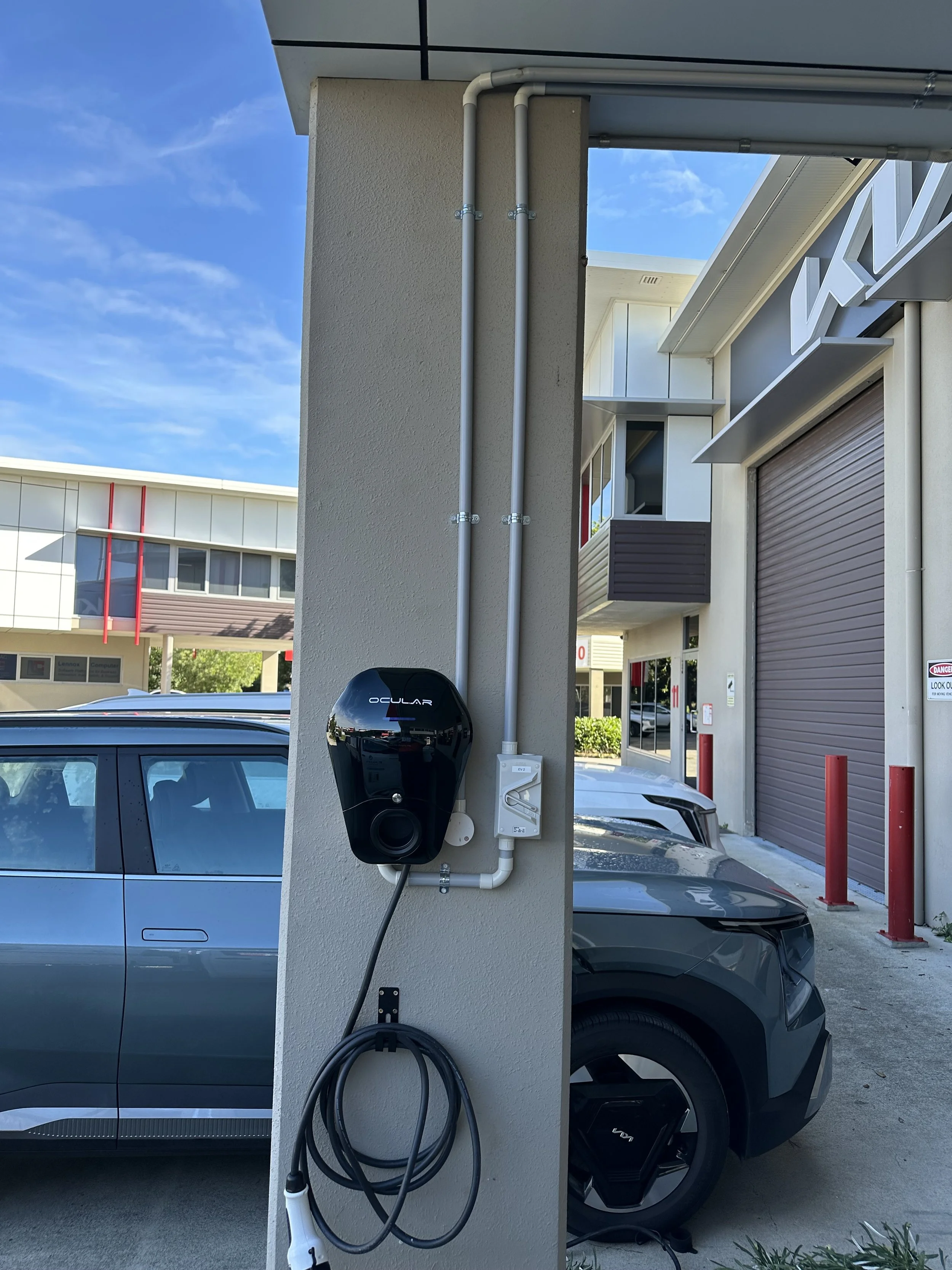 Electric vehicle charging station with a charging cable and a black charging unit attached to a beige pillar, with cars parked in the background outside a commercial building.