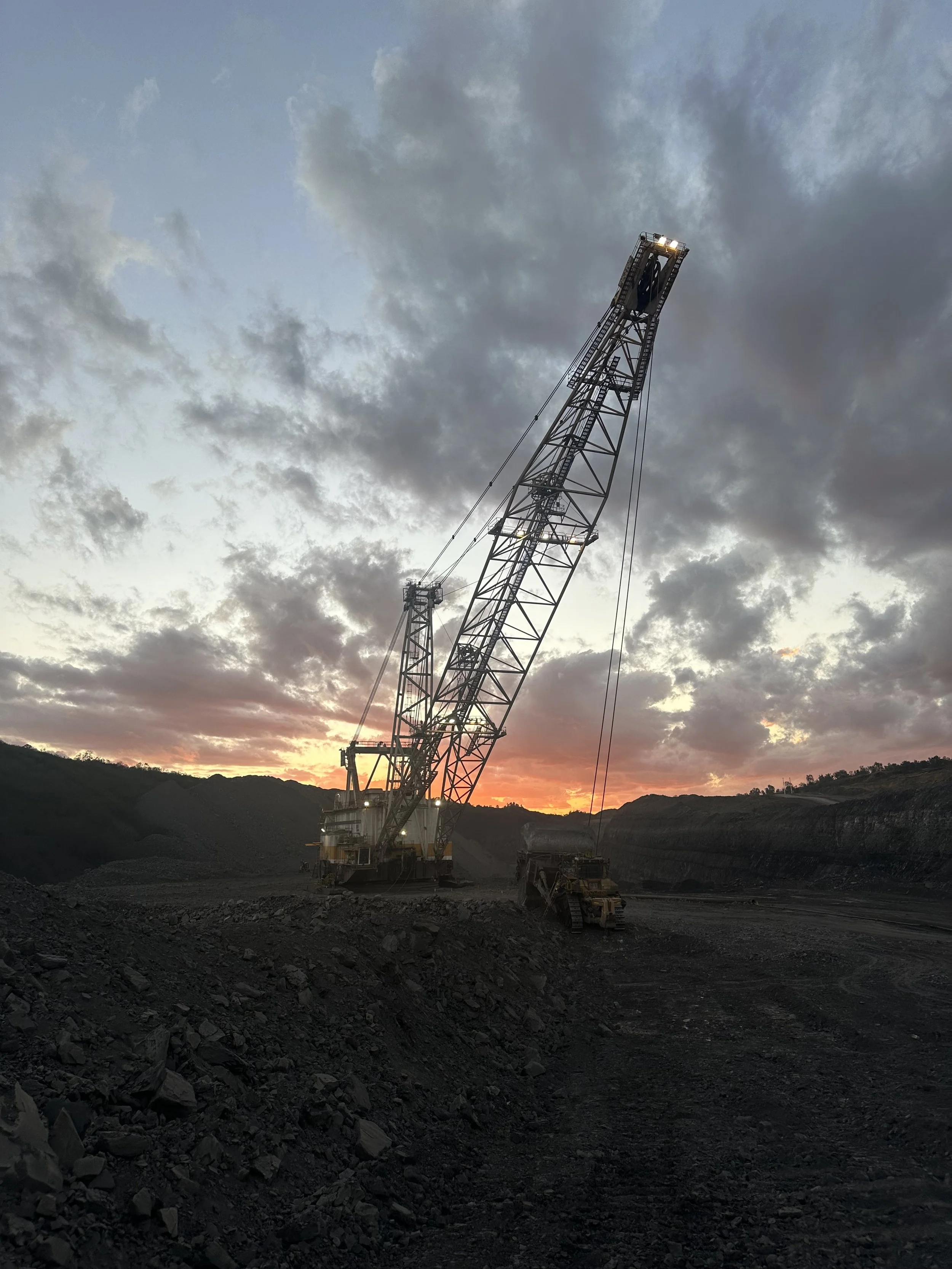 A large construction crane at a mining site during sunset with a cloudy sky.