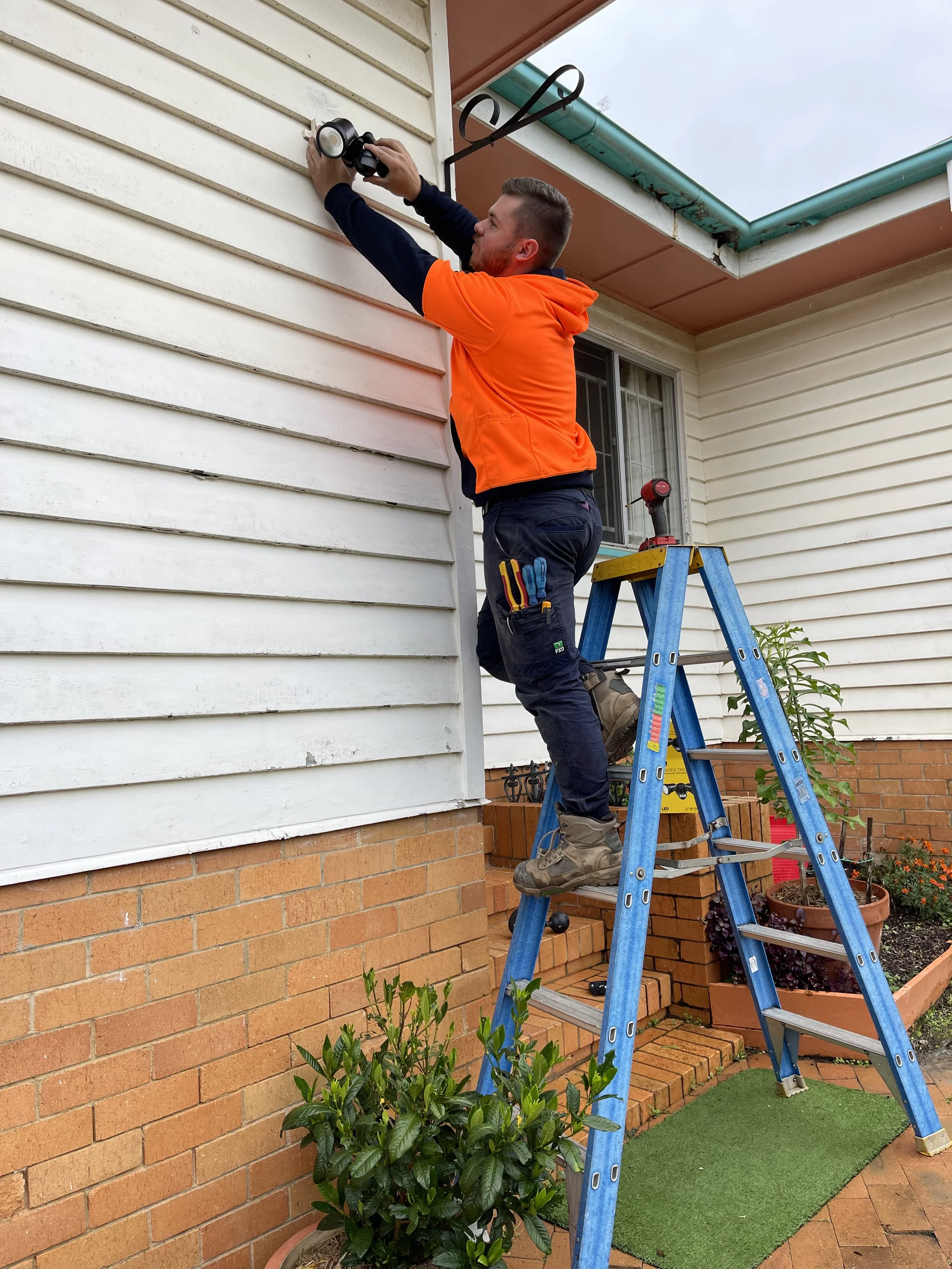 Man standing on a blue ladder fixing or installing a security camera on the exterior wall of a house. The house has white horizontal siding and a brick foundation, with a window and potted plants nearby.