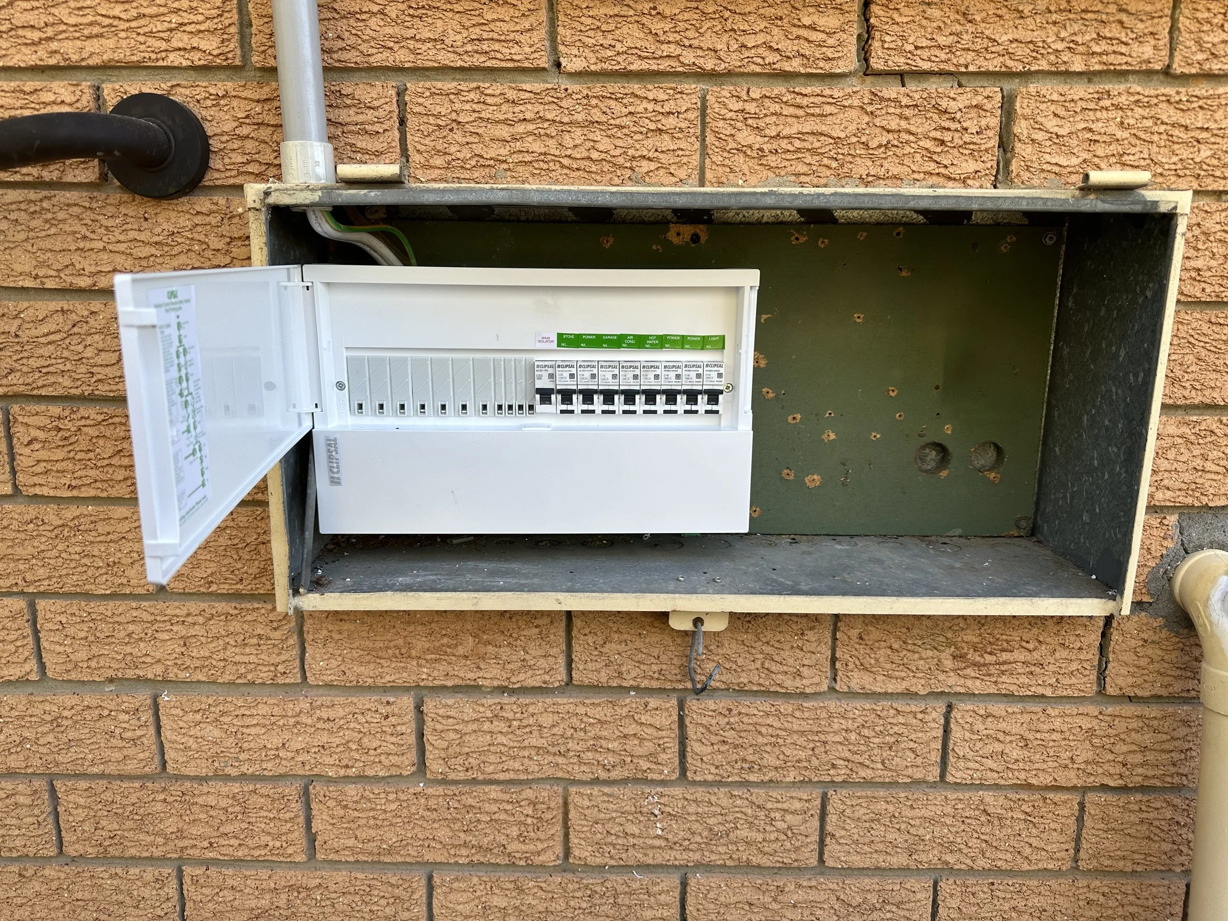 An empty electrical box on a brick wall with exposed wires and a circuit breaker panel inside.