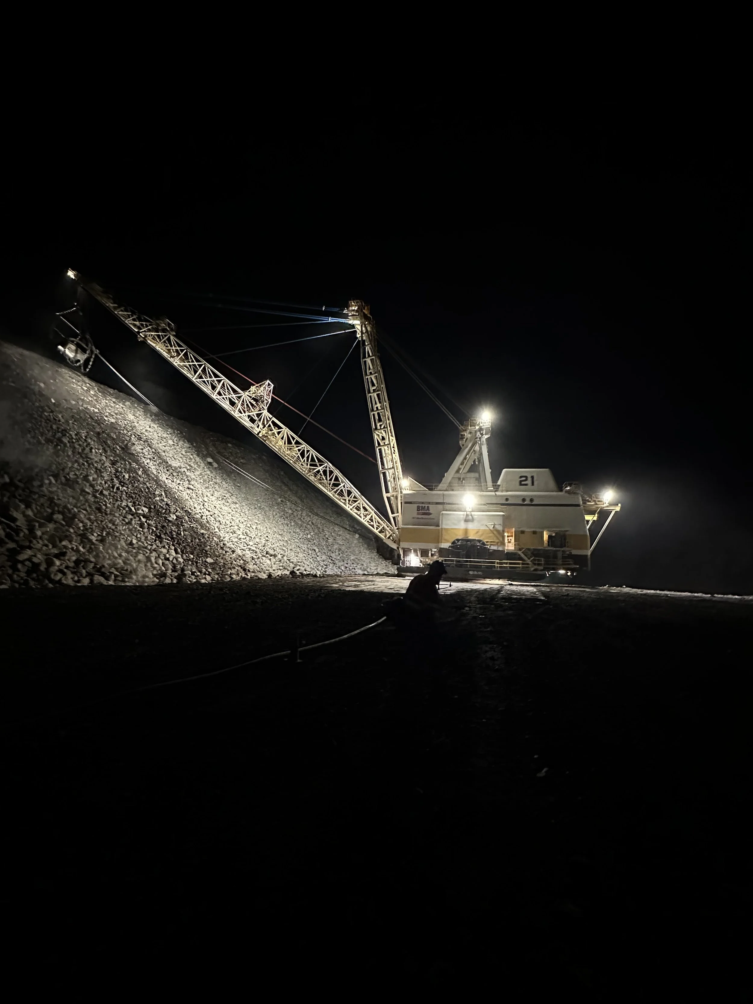 A large construction crane illuminated at night, positioned on a construction site with piles of gravel, with no background visible beyond the dark night sky.