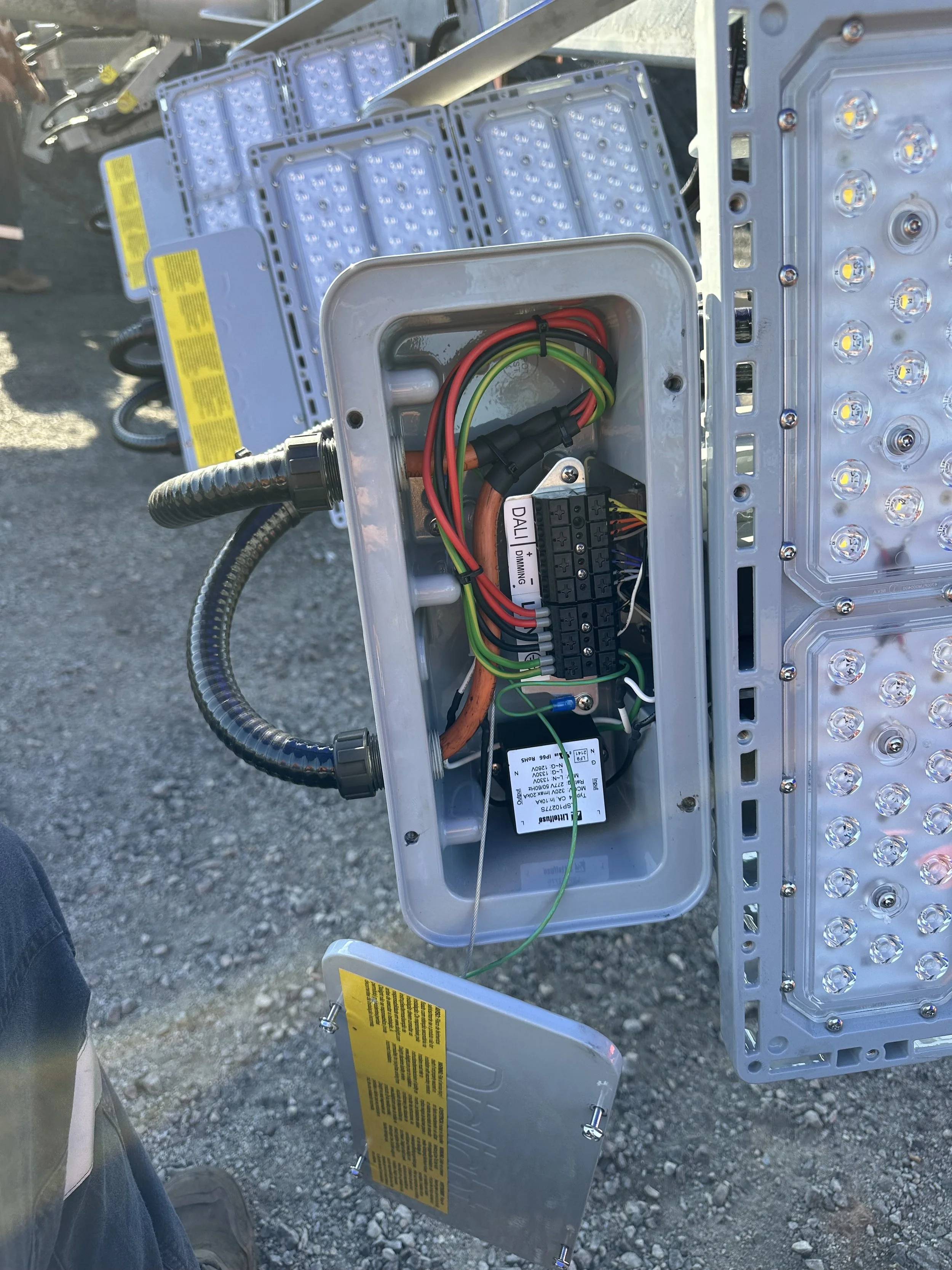 Close-up of an open electrical panel with wiring inside and LED lights on a metal frame, outdoor setting with gravel ground.