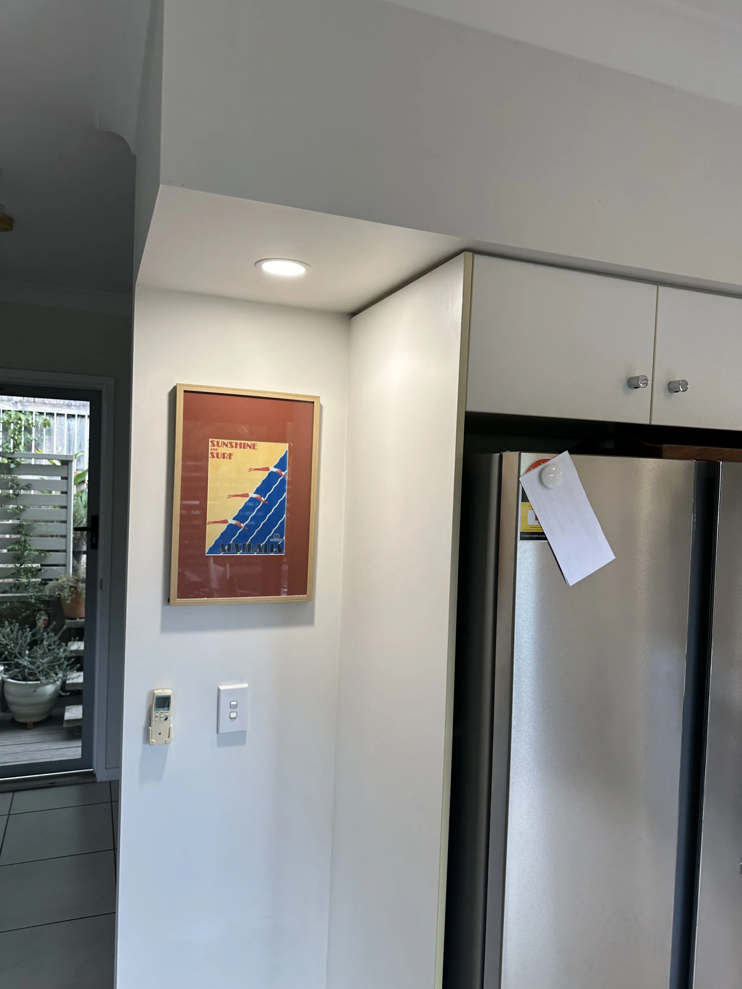 A kitchen wall with a framed poster, a white wall with switches, and a stainless steel refrigerator with a note attached.