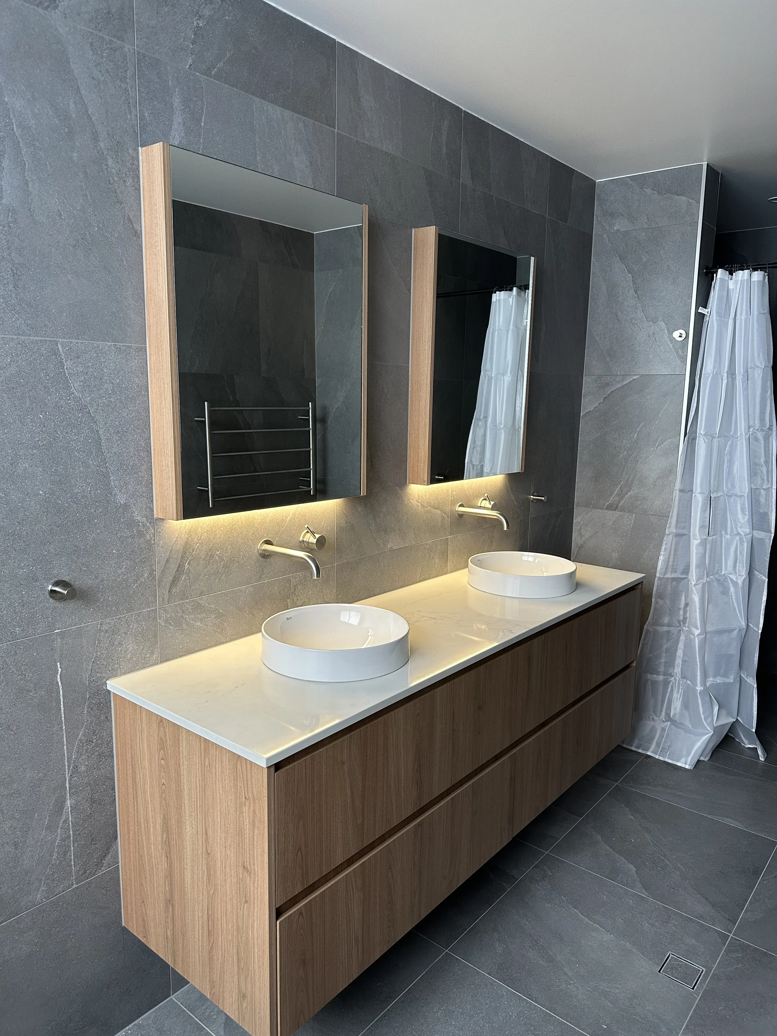 Modern bathroom with dark gray tiled walls and floor, a long wooden vanity with two round white sinks, large mirrors, and a shower area with a white curtain.