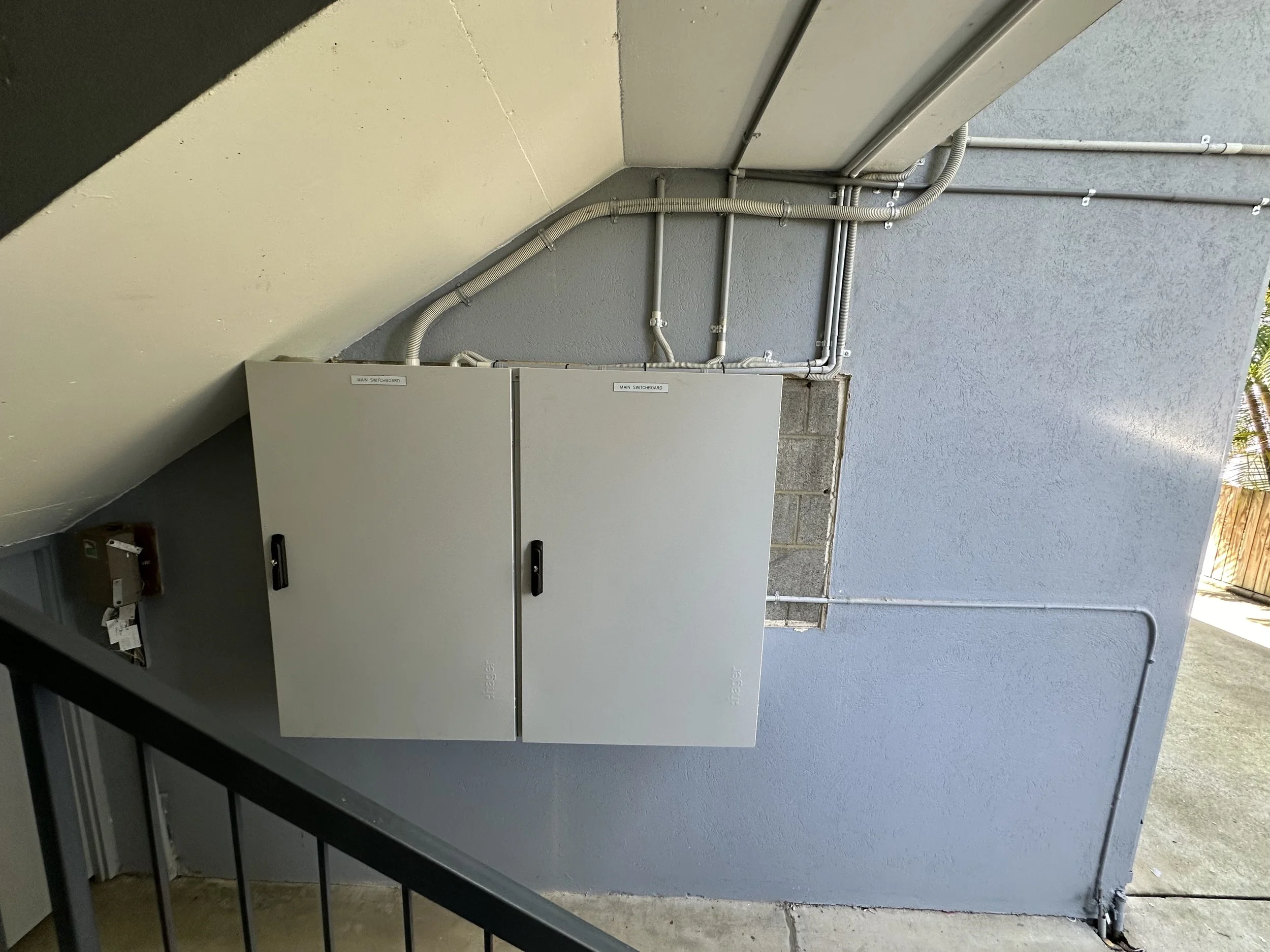 Electrical panel with labeled main switchboard and conduit pipes on a gray exterior wall.