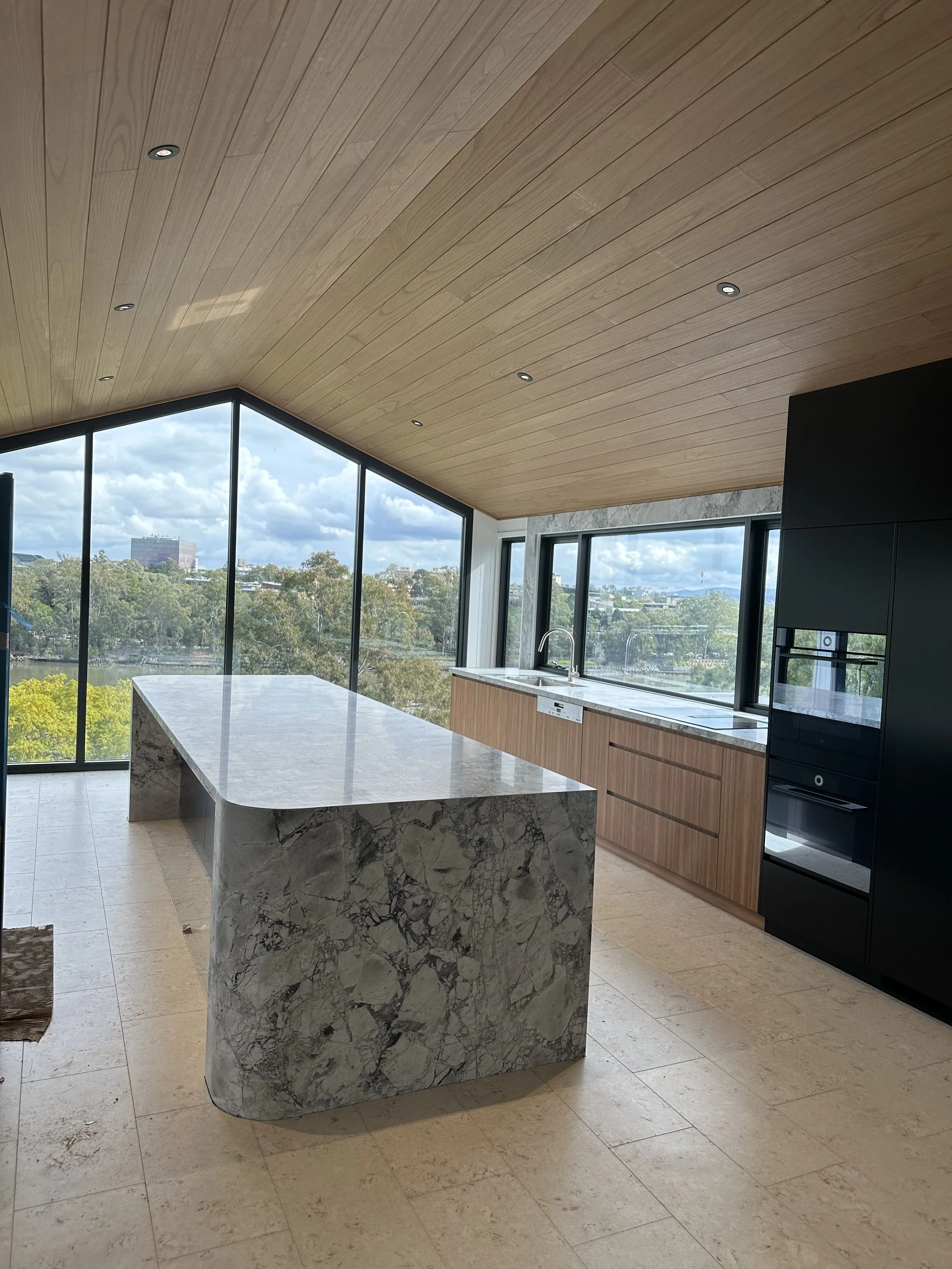 Modern kitchen with large marble island, wooden cabinets, black appliances, and floor-to-ceiling windows showing a cityscape and cloudy sky.