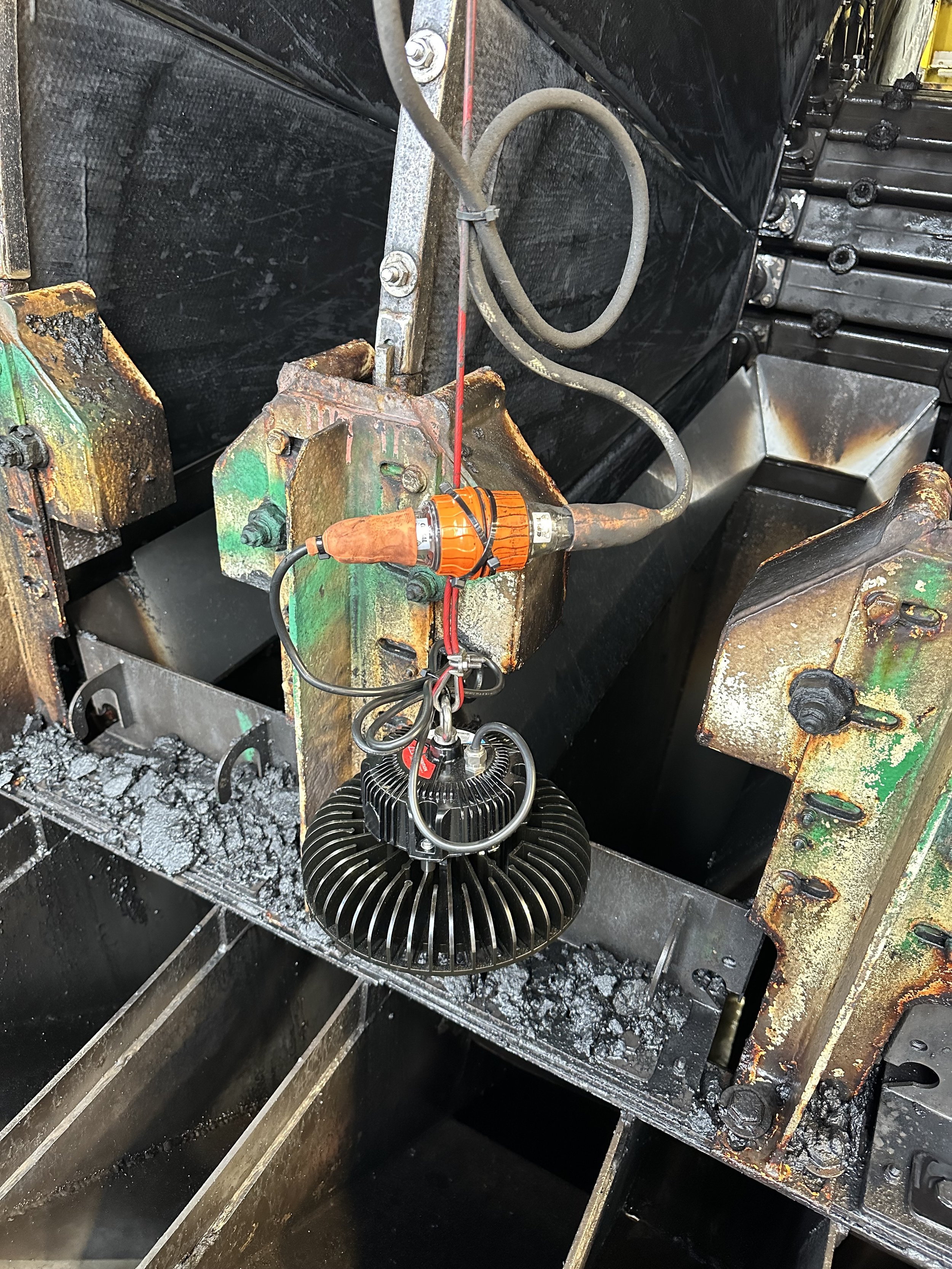 Industrial machinery with a large black container, rusted metal parts, a heating element, and electrical wiring, with a background of charred and soot-covered surfaces.