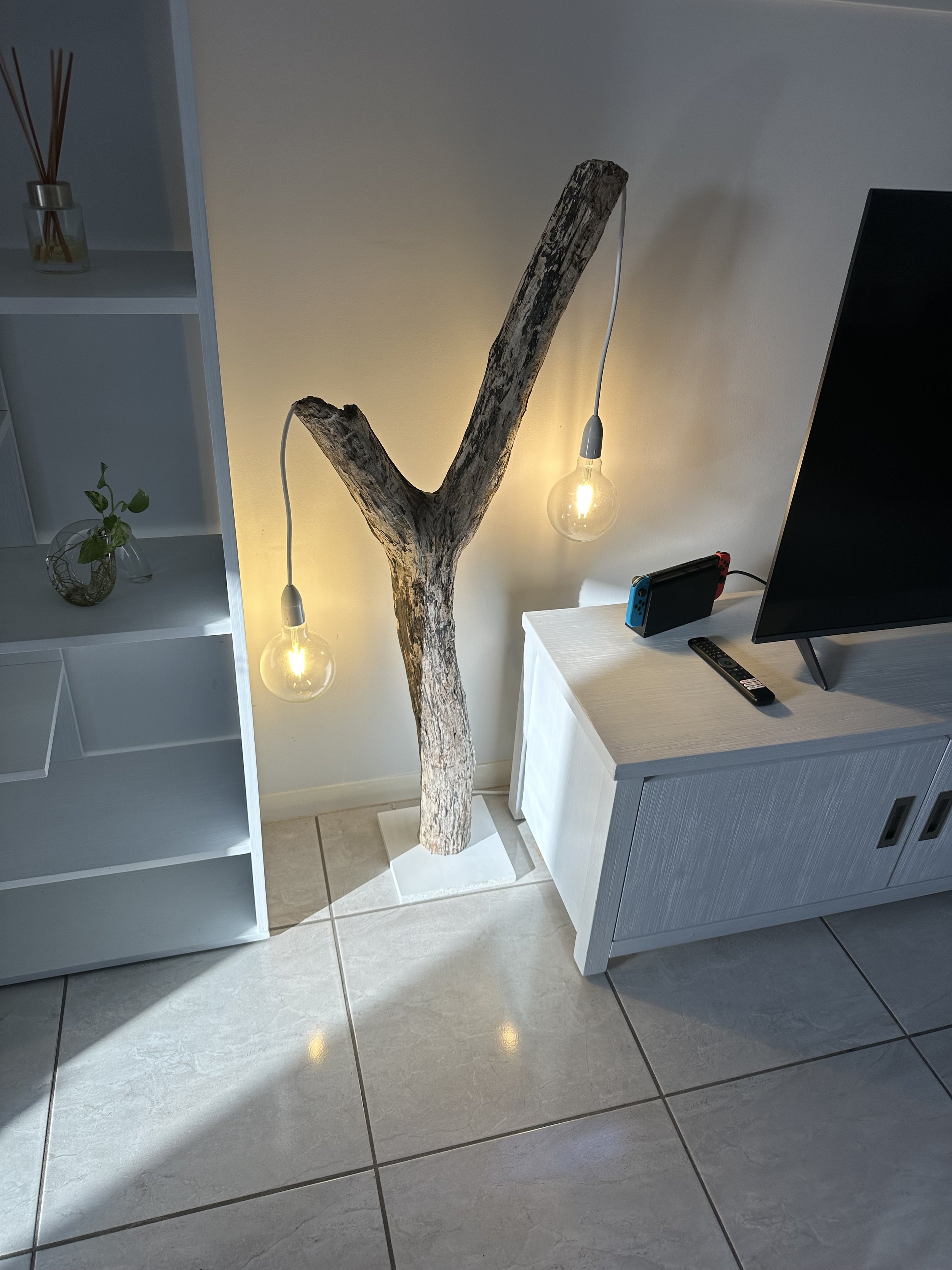 A TV on a white cabinet next to a rustic tree trunk with hanging light bulbs. To the left, a white shelving unit with decorative items including a small plant and a glass jar with wooden sticks.