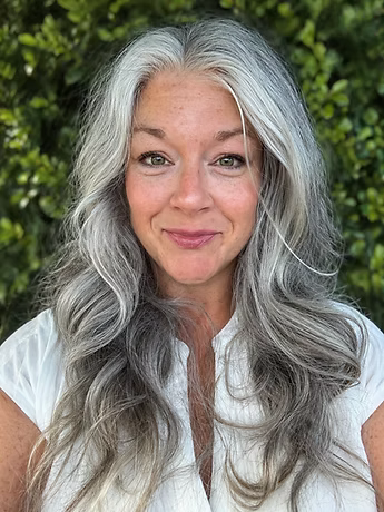 A woman with long, gray hair, smiling, standing outdoors with green foliage in the background.