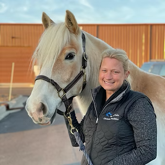 Woman smiling next to a white horse wearing a halter at an outdoor equestrian facility.