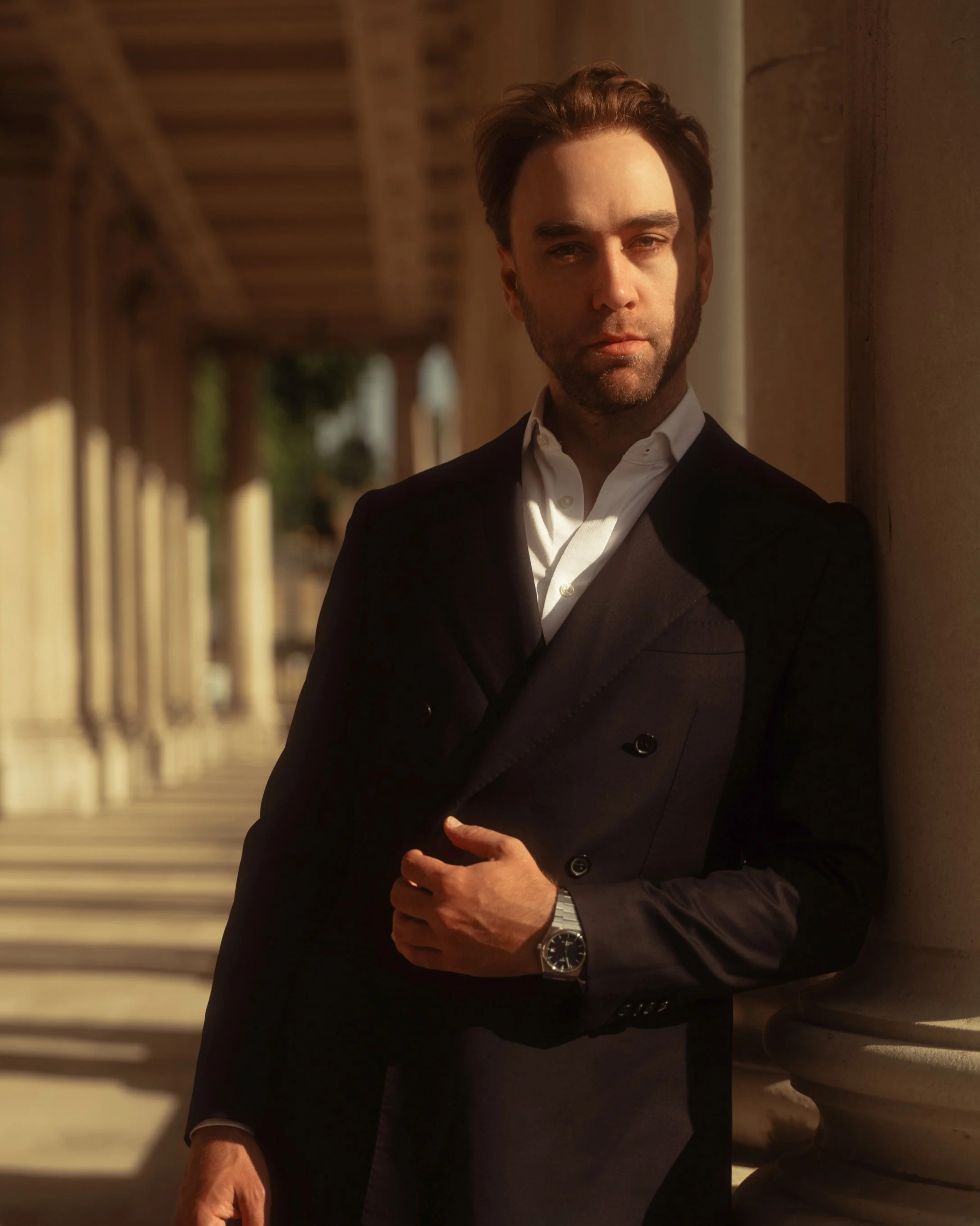 Rian Strauss, Founding Principal Partner of Strauss Partners. A man with dark hair, beard, and light skin wearing a black suit jacket, white dress shirt, and silver watch, standing with arms crossed, leaning against a wall outdoors during daylight.