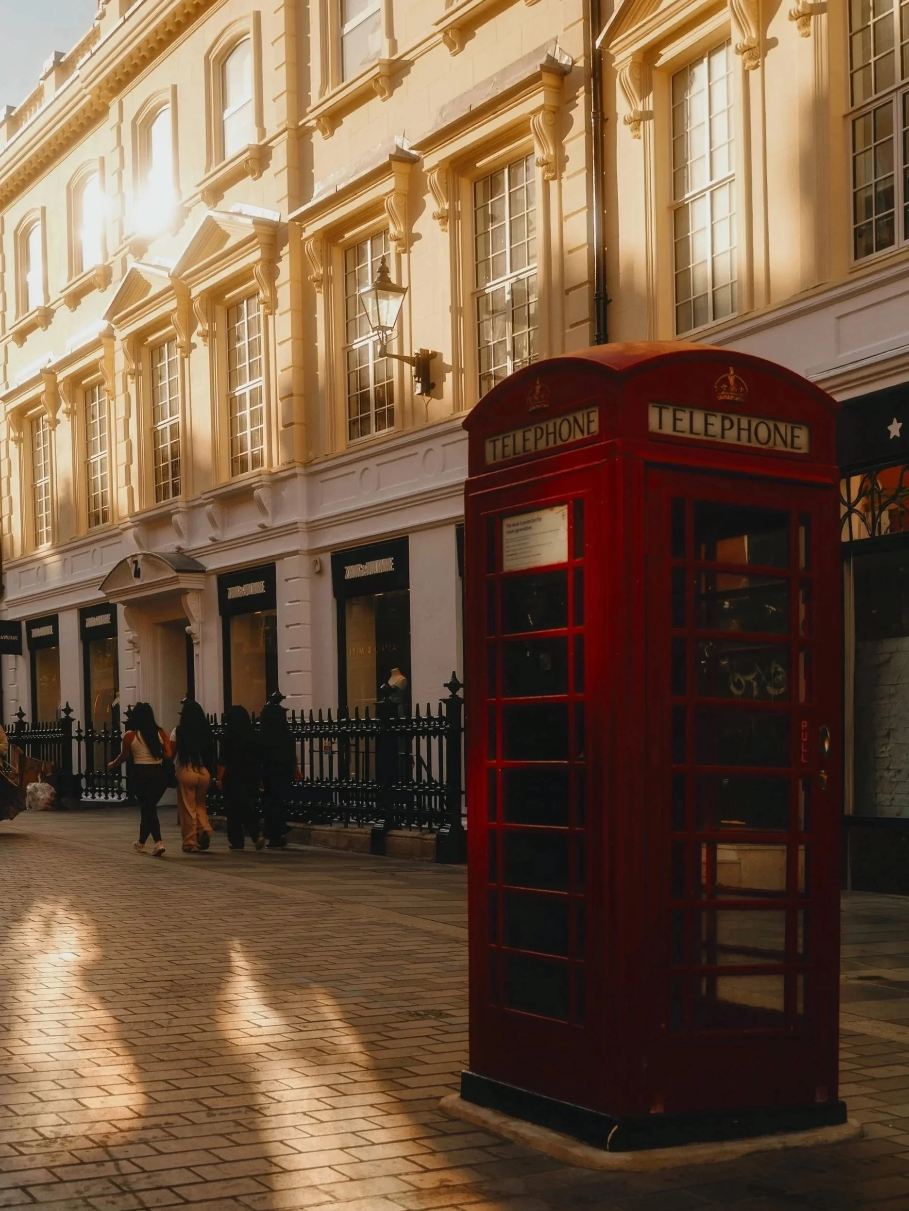 Strauss Partners Contact. Red British telephone booth on a London sidewalk in front of a large light-colored building with multiple windows, some people walking on the street, and sunlight reflecting off the building.