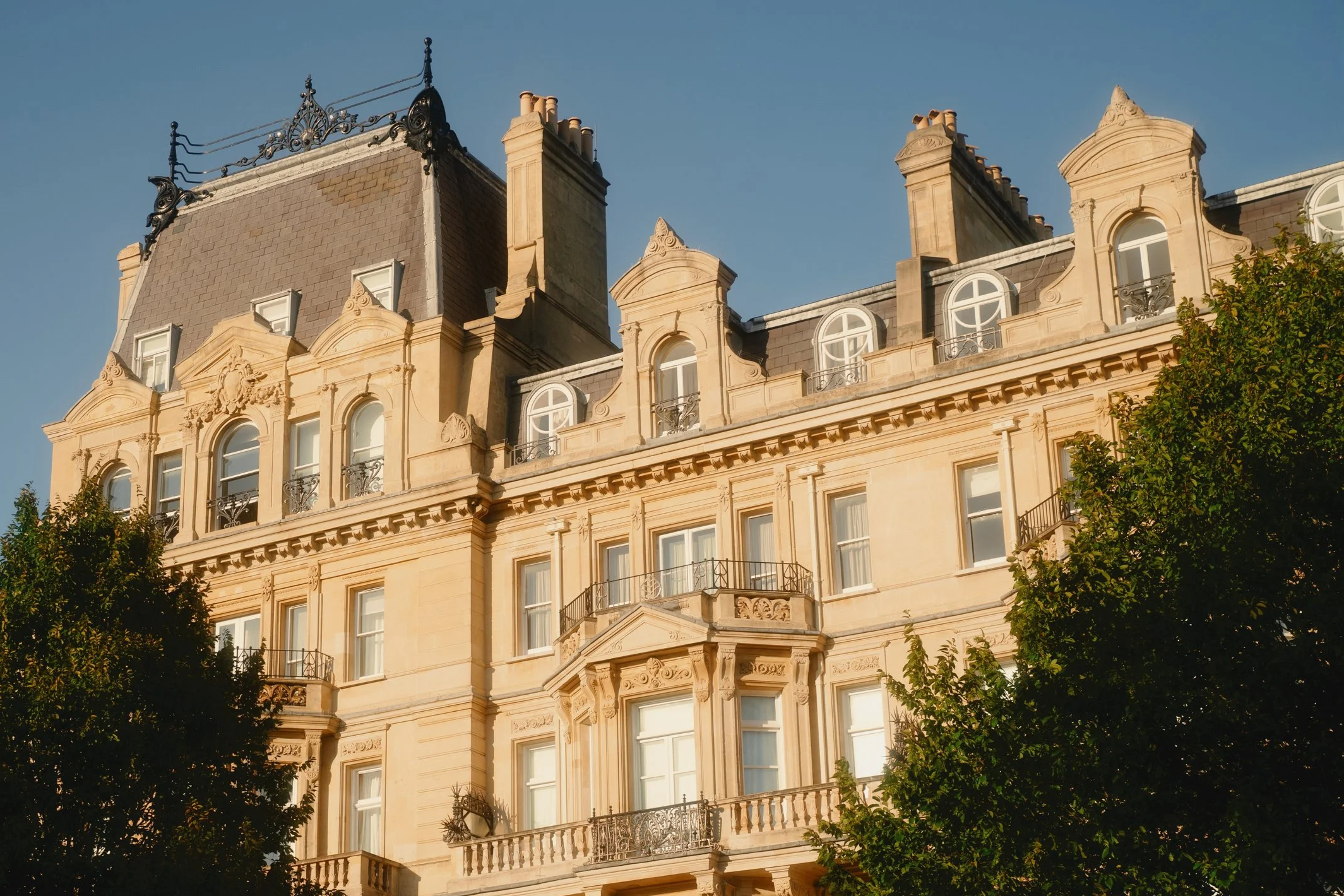 A historic multi-story European building with beige stone facade, decorative moldings, rounded windows, and wrought iron balcony railings, set against a clear blue sky with trees in the foreground, located in Prime London.