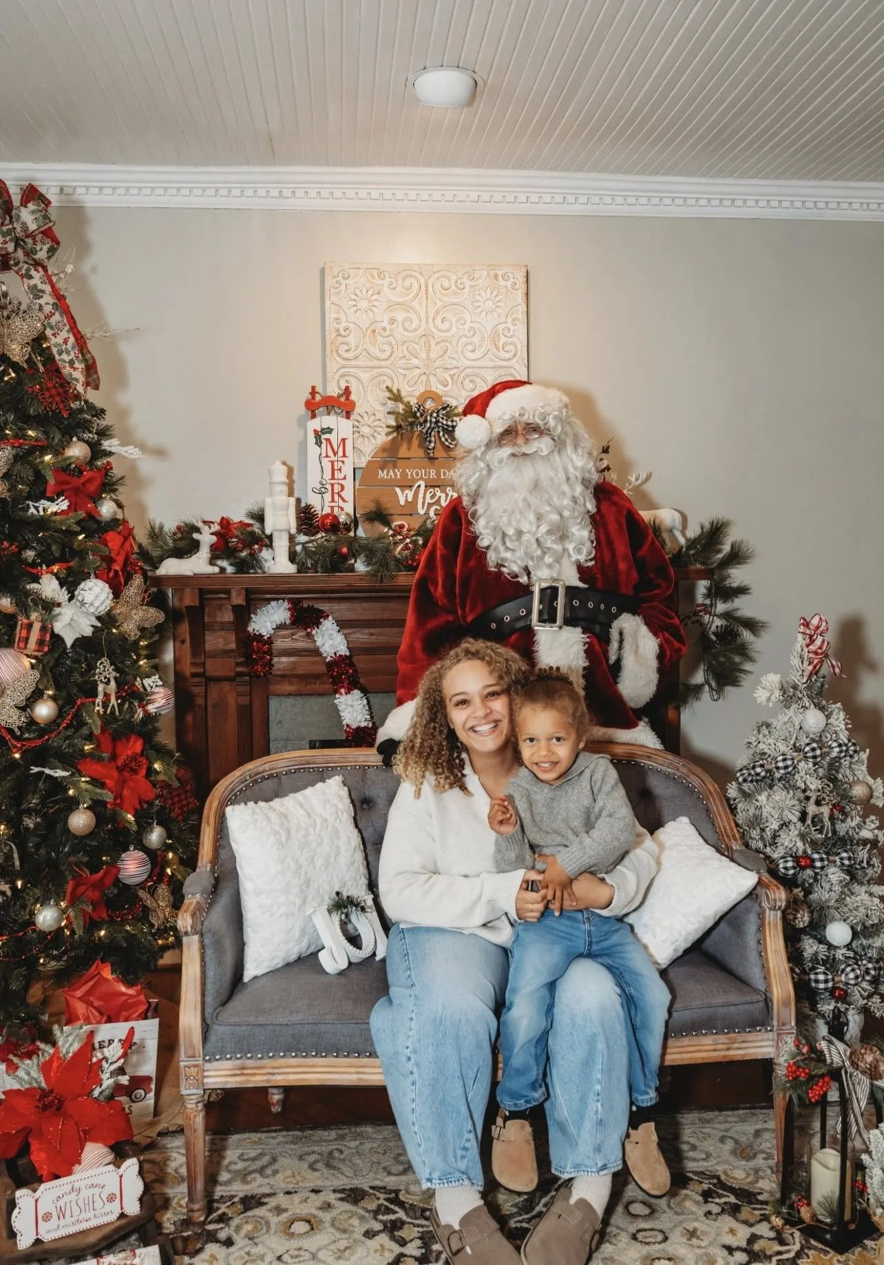 A woman and a young boy sitting on a gray sofa with white pillows, smiling at the camera, with Santa Claus standing behind them. The background is decorated for Christmas with a decorated tree, ornaments, and festive signs.