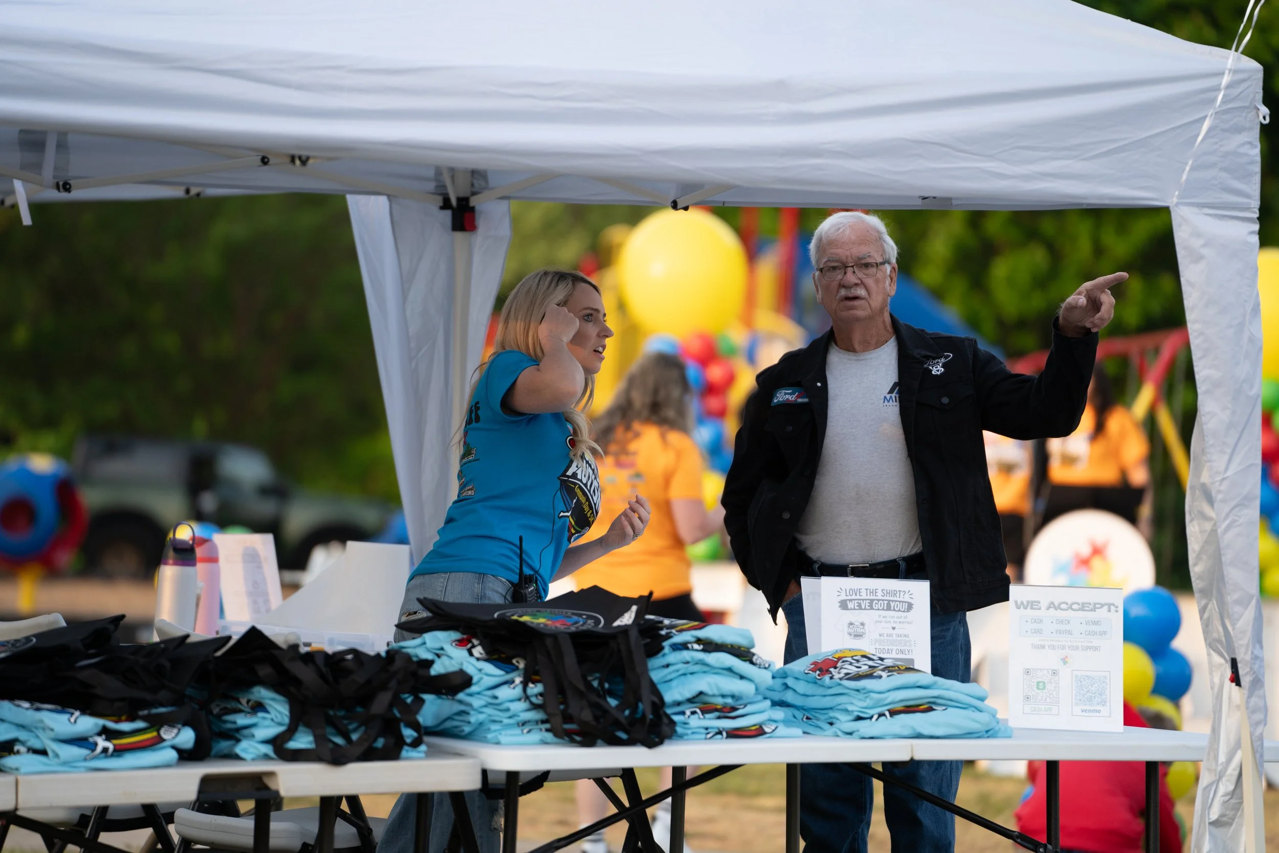 A man and a woman standing at a table with t-shirts at an outdoor event under a white canopy tent. The man is gesturing with his hand while the woman listens attentively.