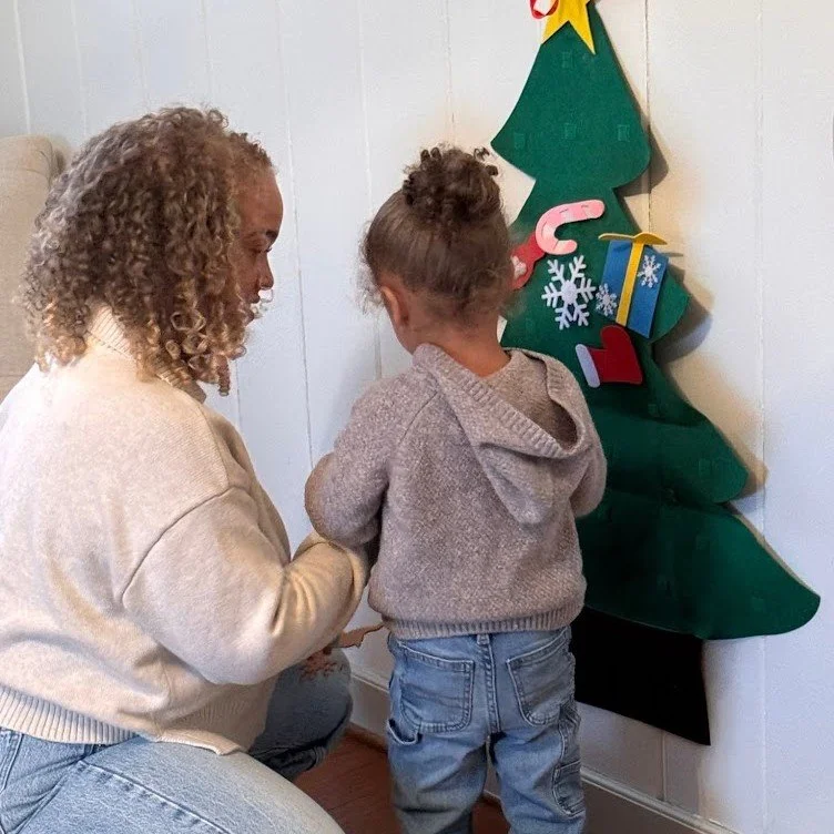 A woman and a young girl with curly hair looking at a decorated felt Christmas tree on the wall, with ornaments including a gift box, candy cane, and snowflakes.