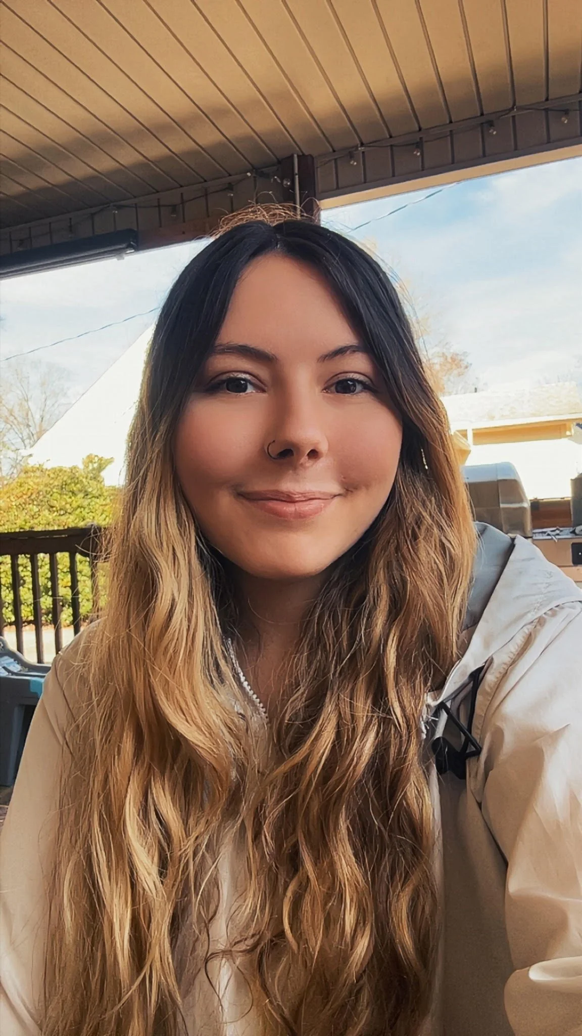 A woman with long, wavy, ombre hair and a nose ring smiling outdoors under a covered patio, with trees and a blue sky in the background.