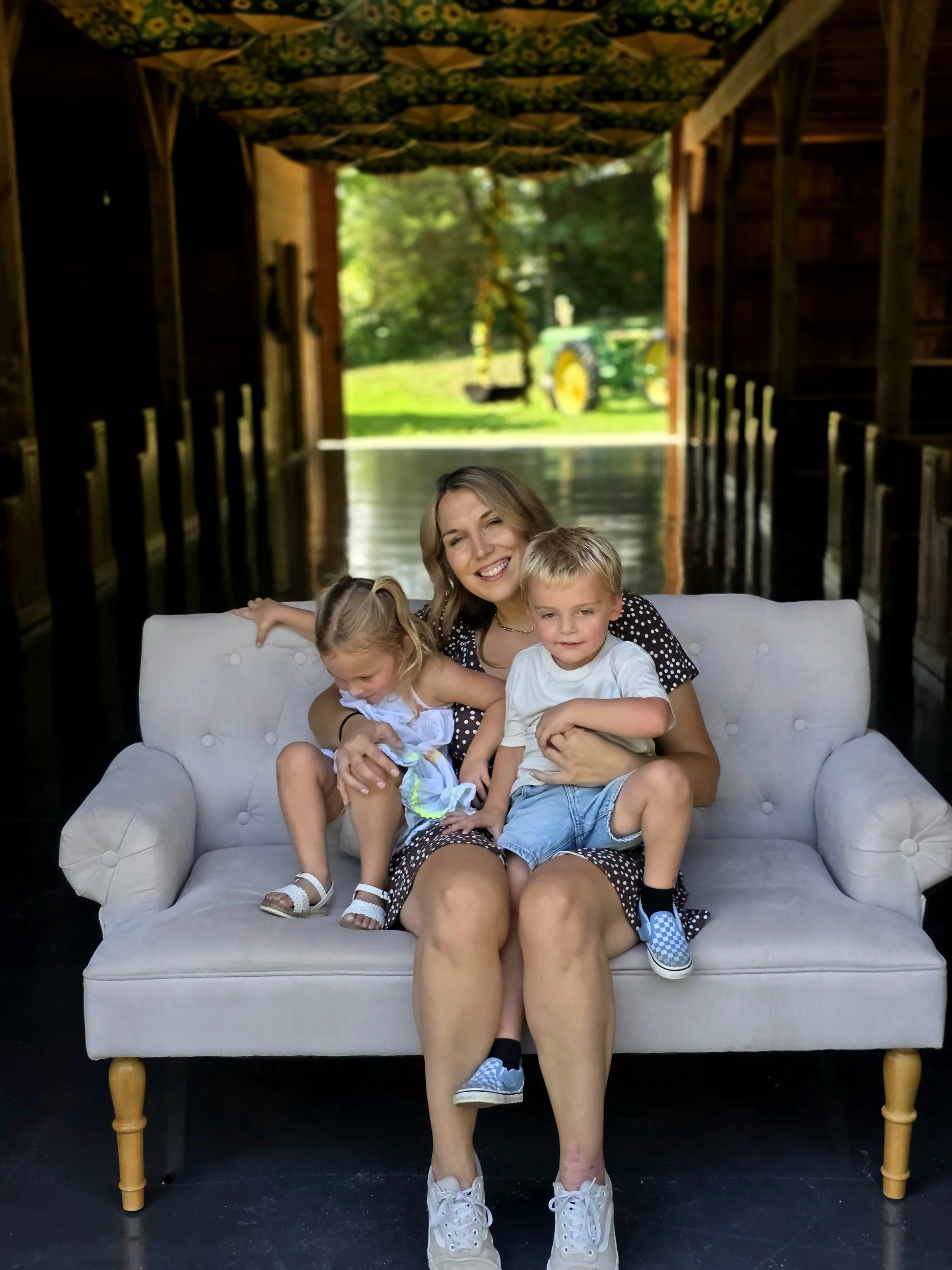 A woman with three children sitting on a white couch inside a barn. Behind them, open doors reveal a green tractor, grass, and trees.