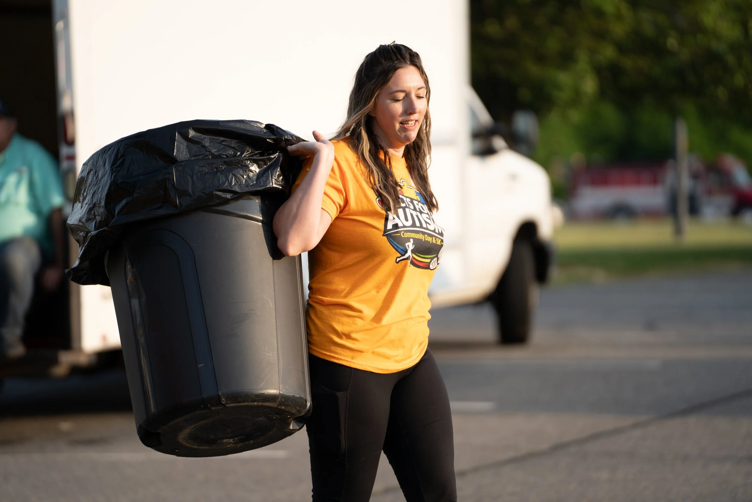A woman wearing an orange t-shirt with a logo, carrying a large black trash bag on her shoulder, walking outside during daylight.