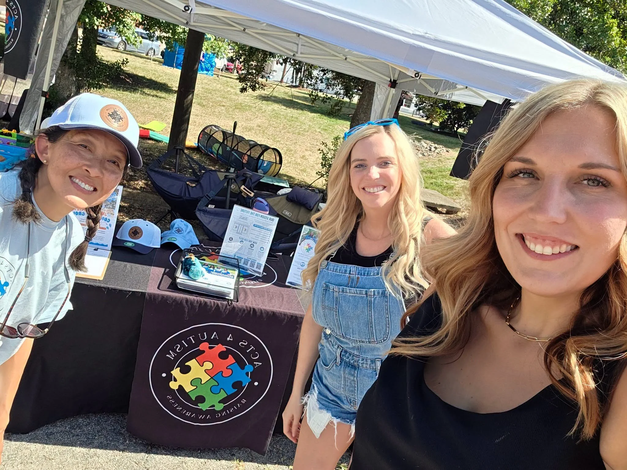 Three smiling women taking a selfie outdoors during a community event or fair, with a table displaying colorful informational materials and merchandise under a white canopy tent.