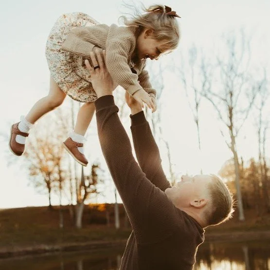 A man lifting a young girl in the air outdoors during sunset, with trees in the background.