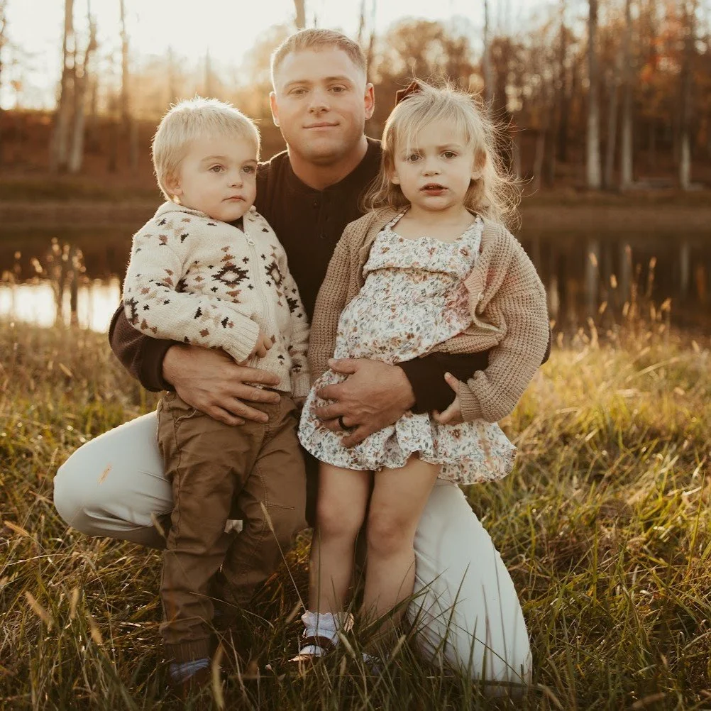 A man with two young children outdoors near a lake at sunset, holding and posing for a photo.