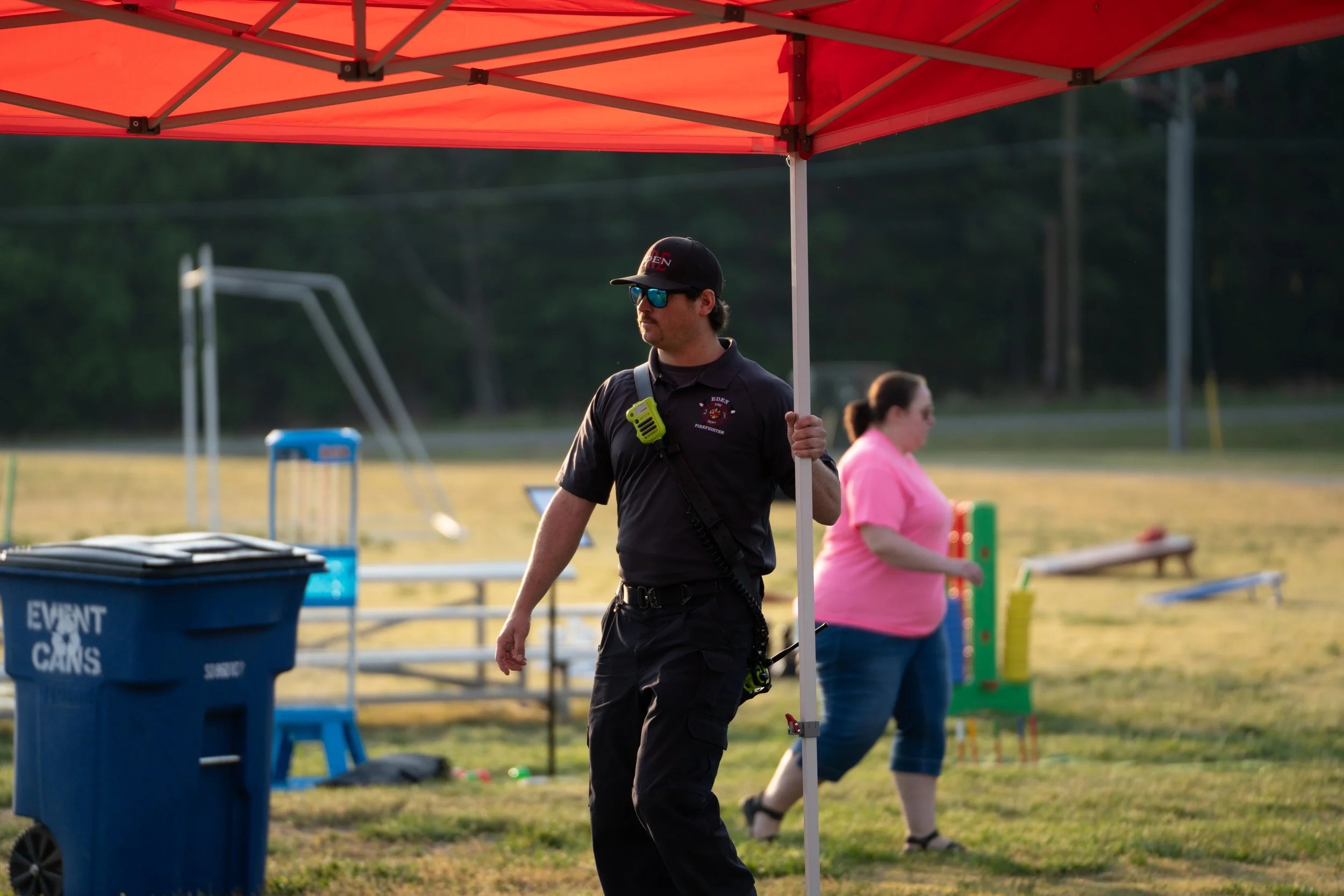A man in firefighter uniform holding a pole under a red canopy at an outdoor event. A woman in pink shirt is in the background. Various equipment and trash bins are visible in the background.