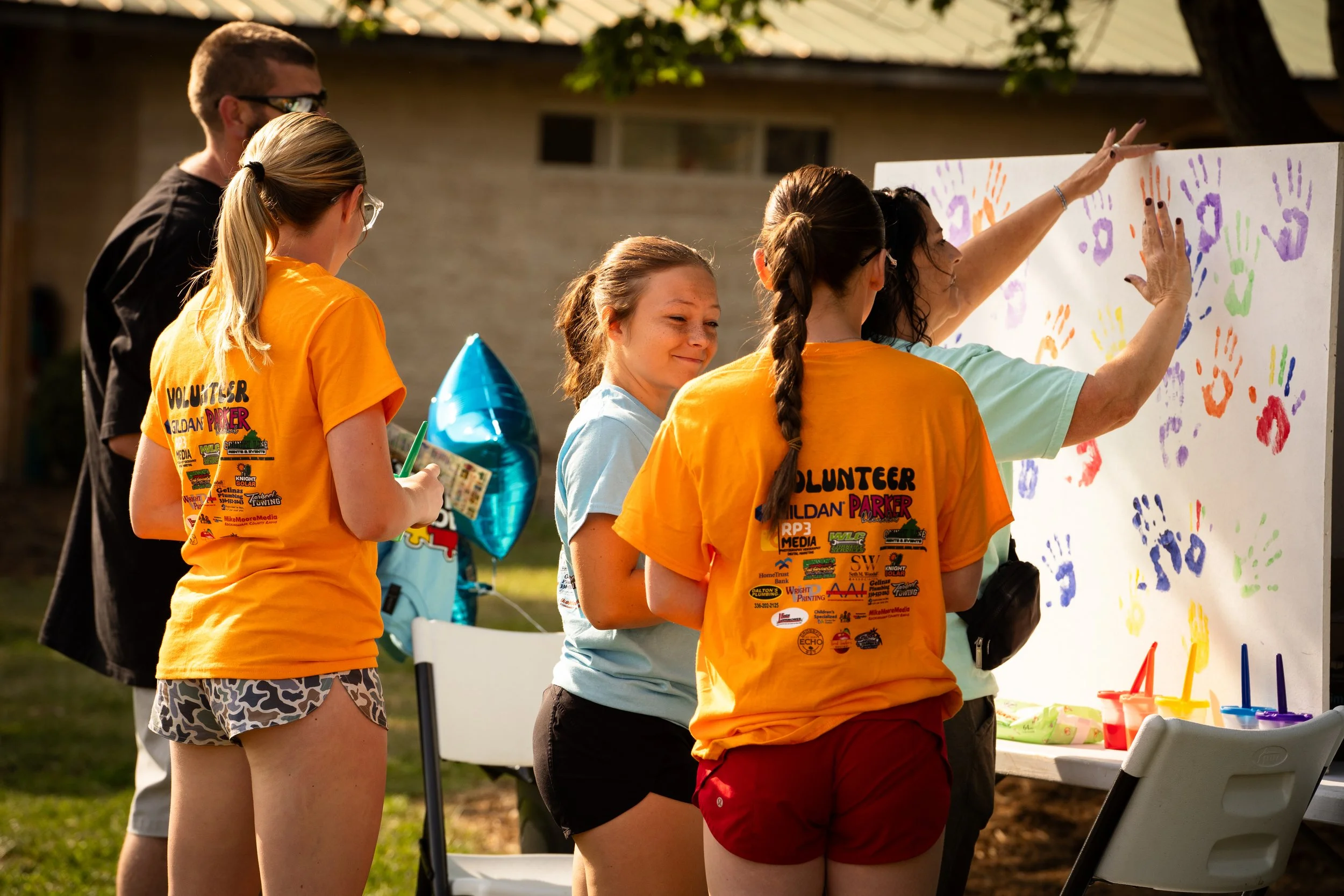 Group of young volunteers at an outdoor event, decorating a large white board with colorful handprints and numbers. They are wearing orange volunteer shirts and are surrounded by tables with art supplies.