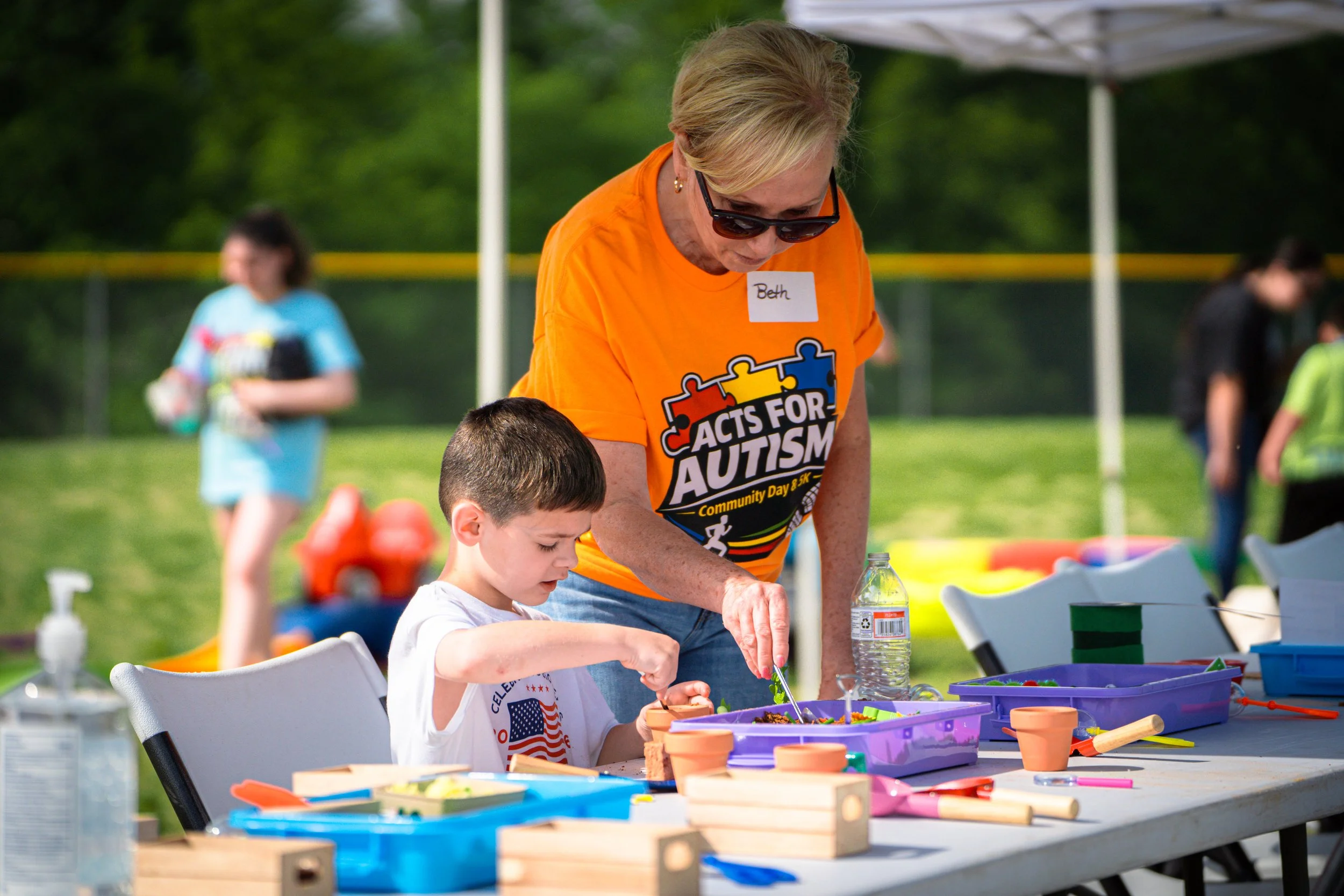 A woman with blonde hair, wearing sunglasses and an orange t-shirt with the words 'Acts for Autism,' assists a young boy at an outdoor event with arts and crafts. There are tables with colorful craft supplies and other children and adults in the background.