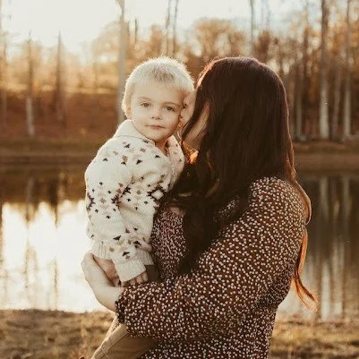 A woman holding a young child outdoors near a pond during autumn, with trees in the background and warm sunlight.