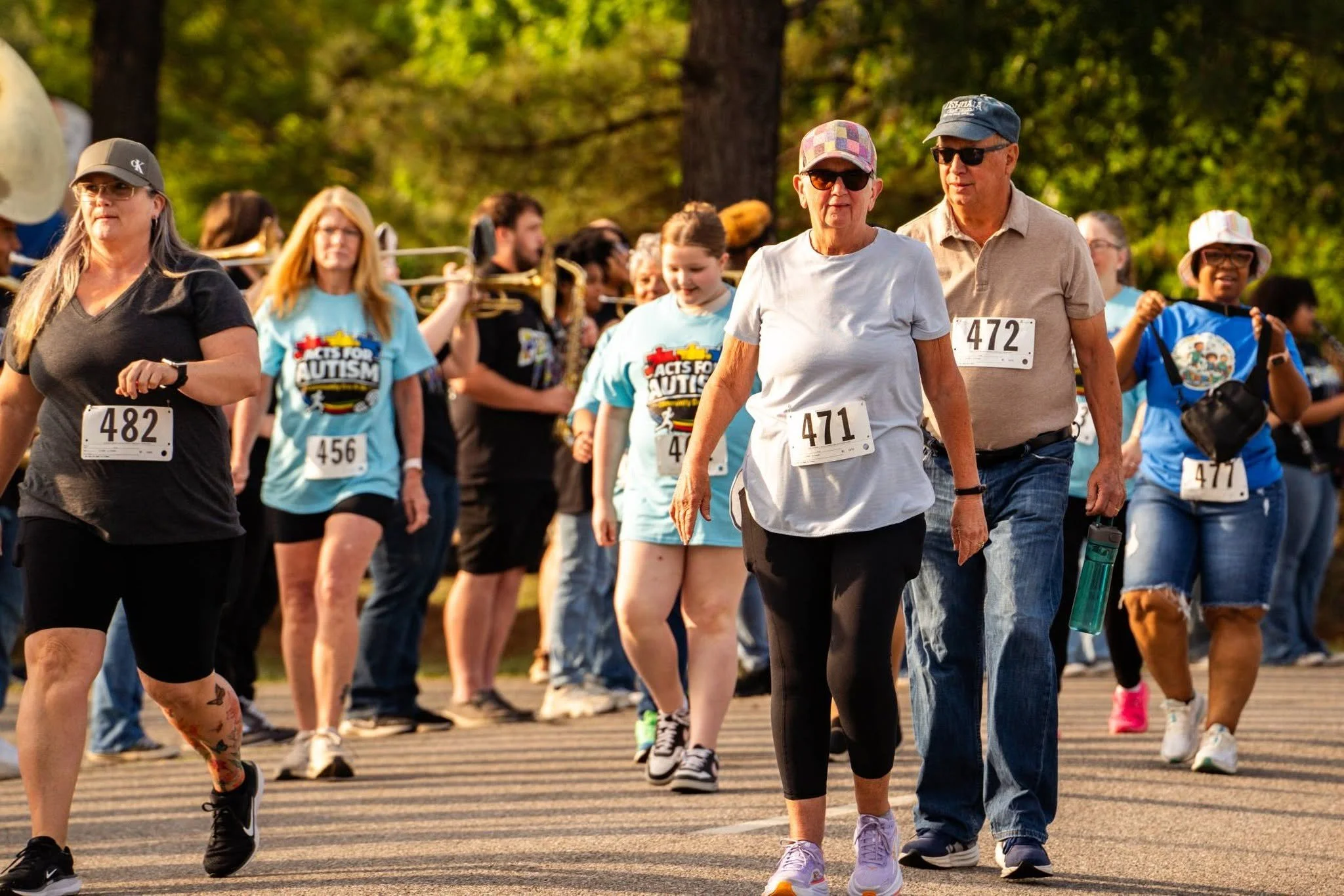 People participating in a charity walk or run event, some wearing bibs with numbers, outdoors in a park with trees in the background.