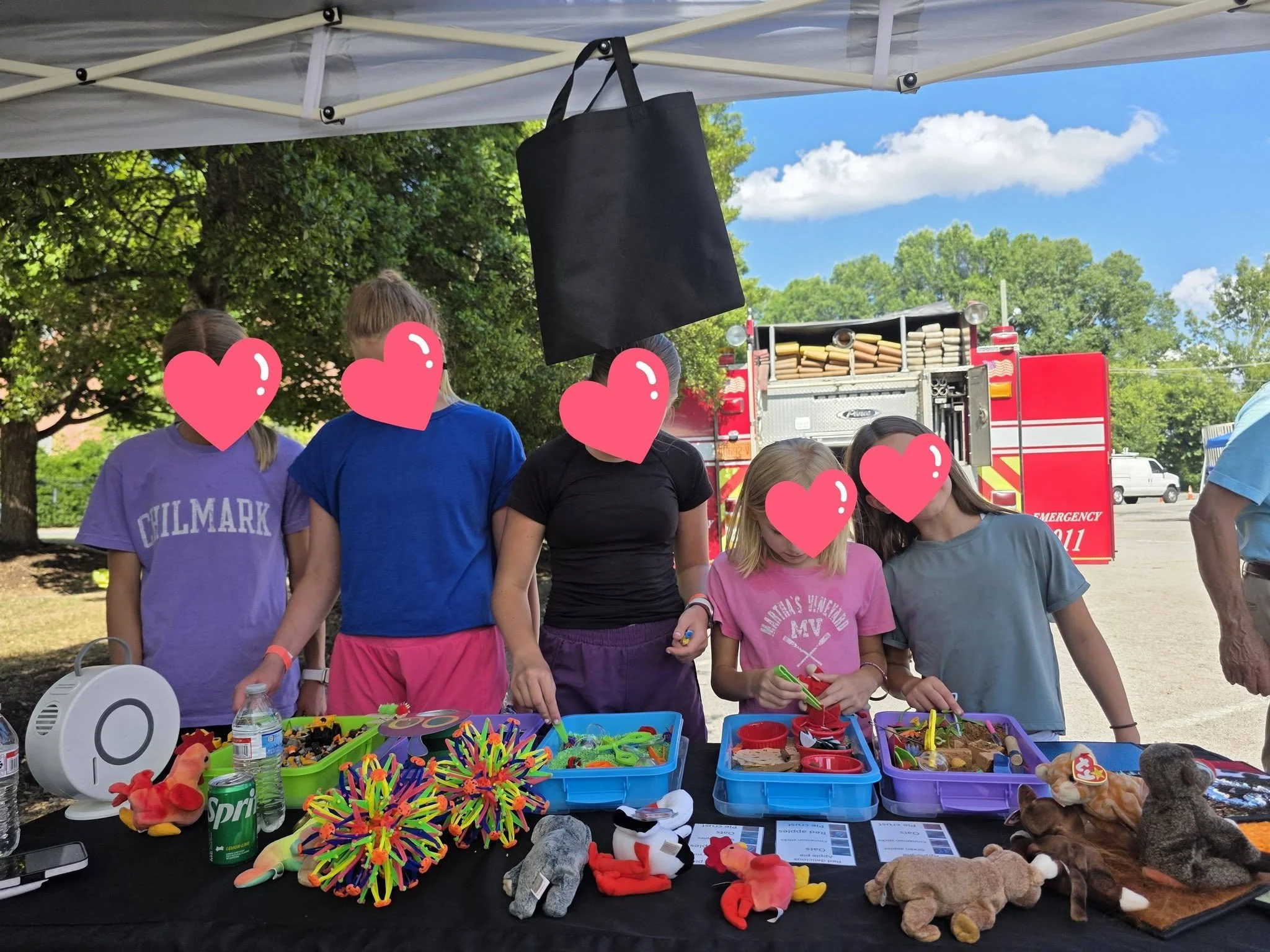 Children at an outdoor toy sale or event, with a table displaying various toys, plush animals, and colorful items under a tent, with a fire truck in the background and trees against a blue sky.