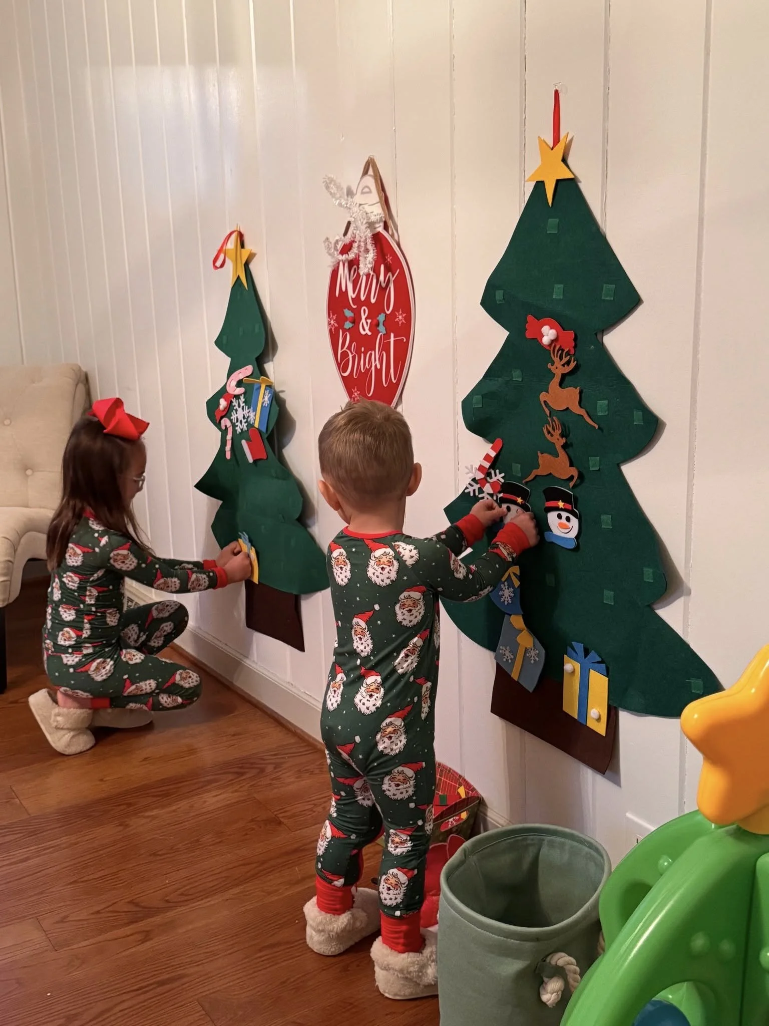 Three children in Christmas pajamas decorating a wall with Christmas trees made of paper and felt, along with holiday decorations. Two children are kneeling, and one is standing, reaching to place or adjust a decoration on the paper Christmas tree. There is a Thanksgiving or Christmas sign on the wall that says 'Merry & Bright' with seasonal decorations.