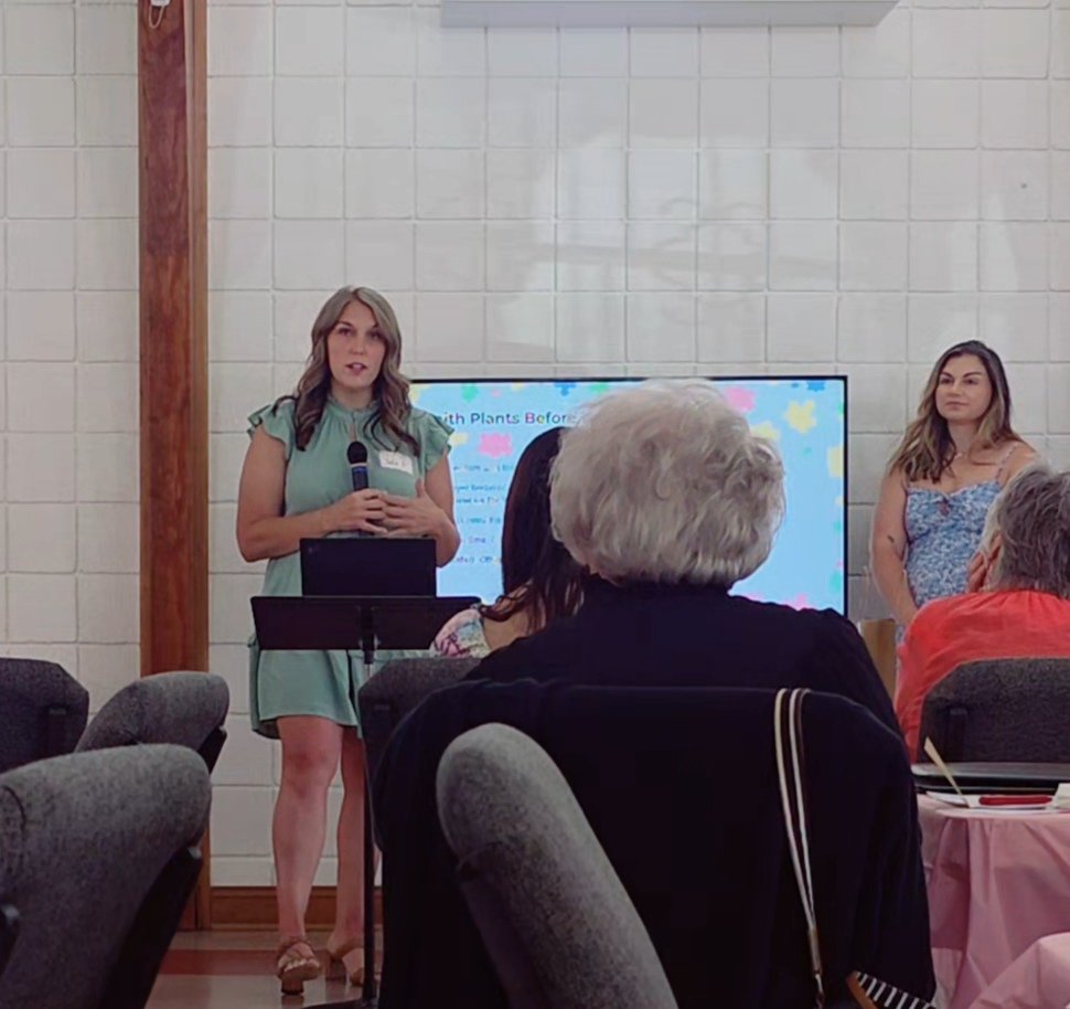 A woman in a green dress holding a microphone speaks in front of a group at a presentation, with a large screen displaying a colorful slide behind her. Another woman stands to the side, and the audience members are seated facing her.
