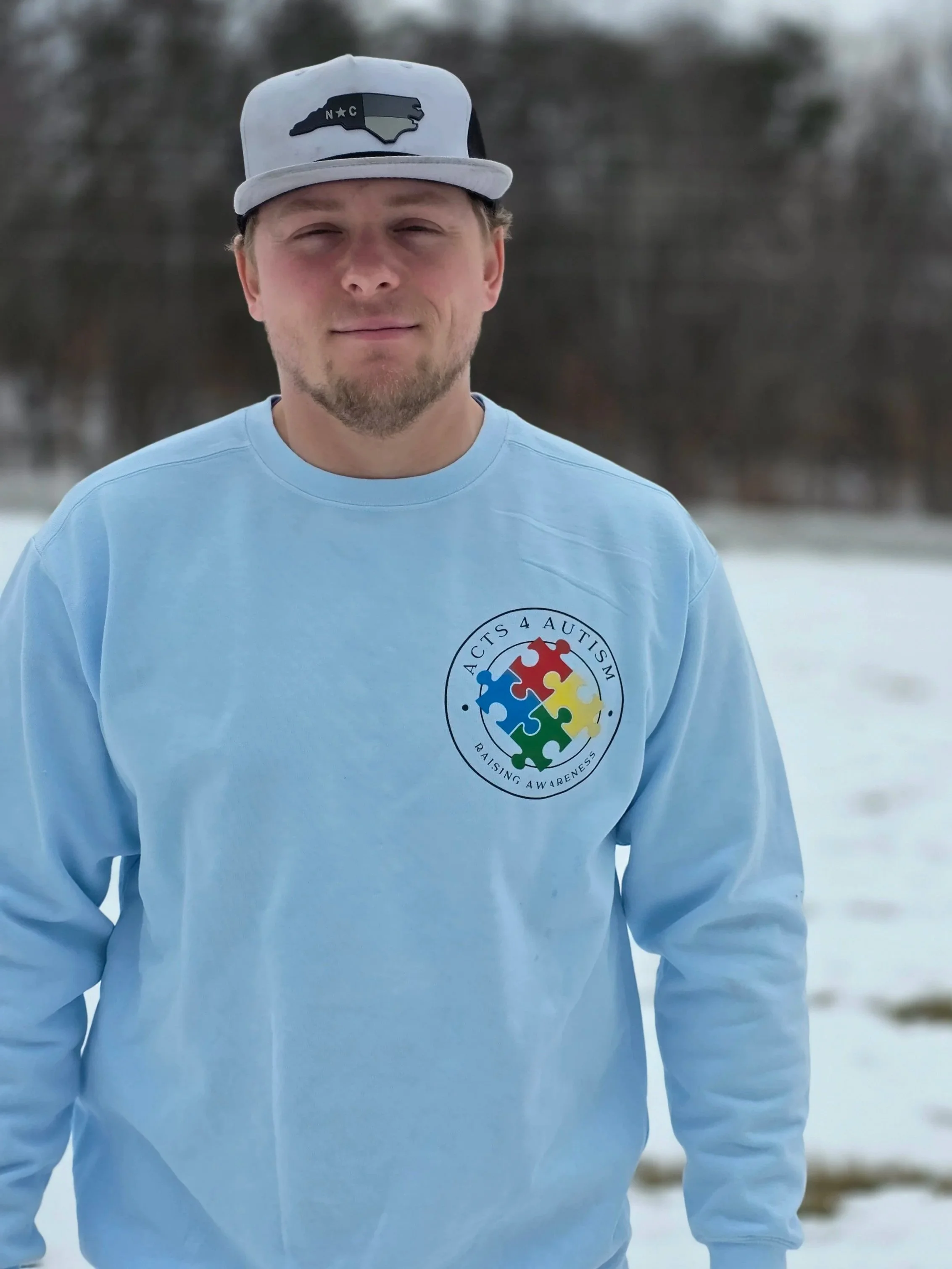 A young man standing outdoors in a snowy landscape, wearing a gray baseball cap with a North Carolina map and star patch, and a blue sweatshirt with a colorful autism awareness puzzle piece logo.