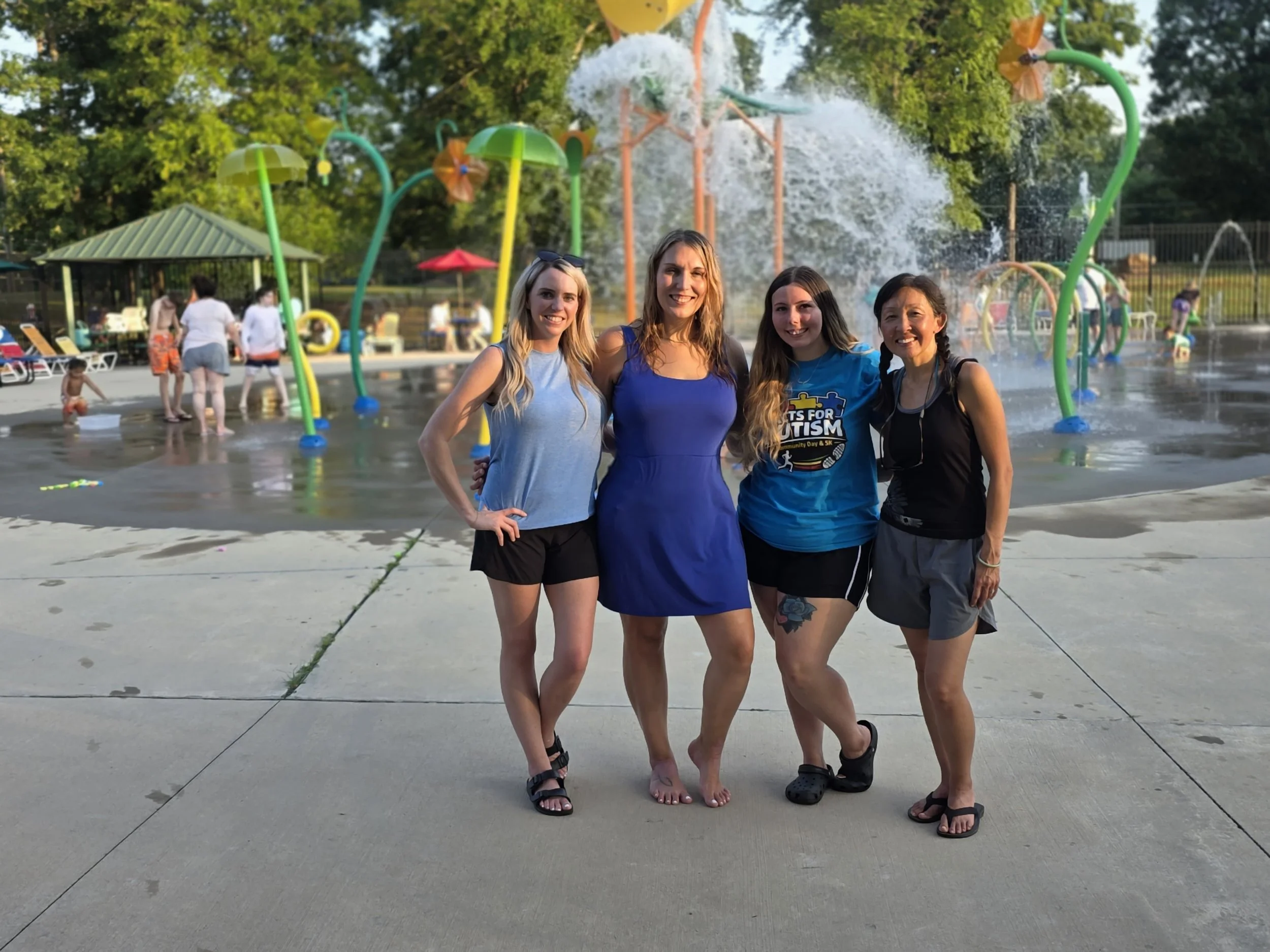 Four women standing together at a splash pad with colorful water sprinklers and children playing in the background, greenery, and a picnic area.