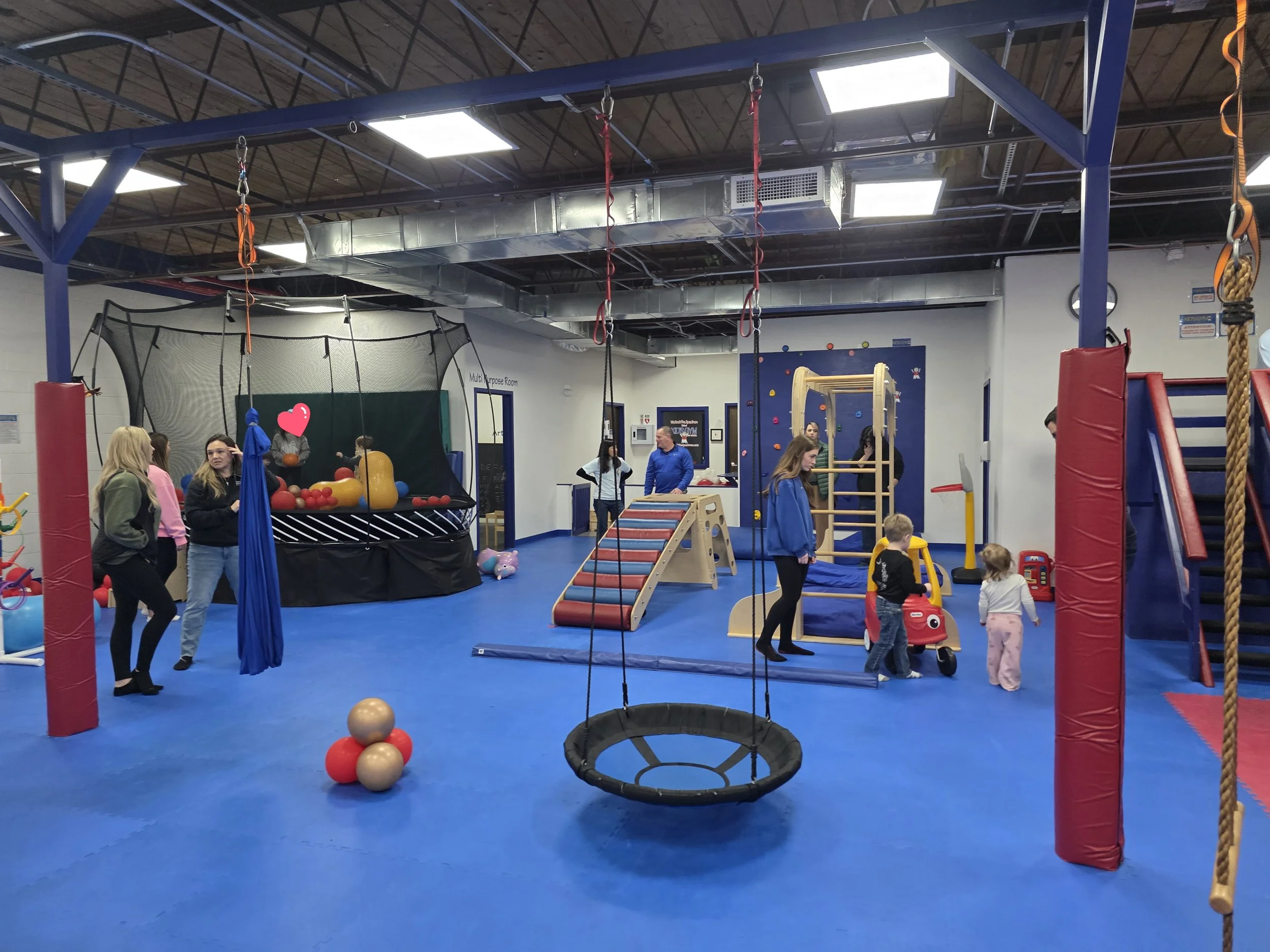 Indoor children's play and activity area with climbing structures, a trampoline, hanging swings, and soft play equipment, populated with children and adults.