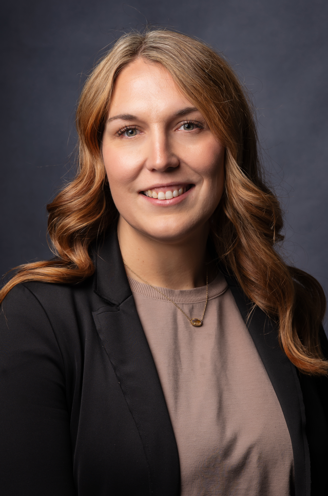 A young woman with long, wavy, light brown hair, smiling in front of a dark gray background. She is wearing a beige top, a black blazer, and a gold necklace.