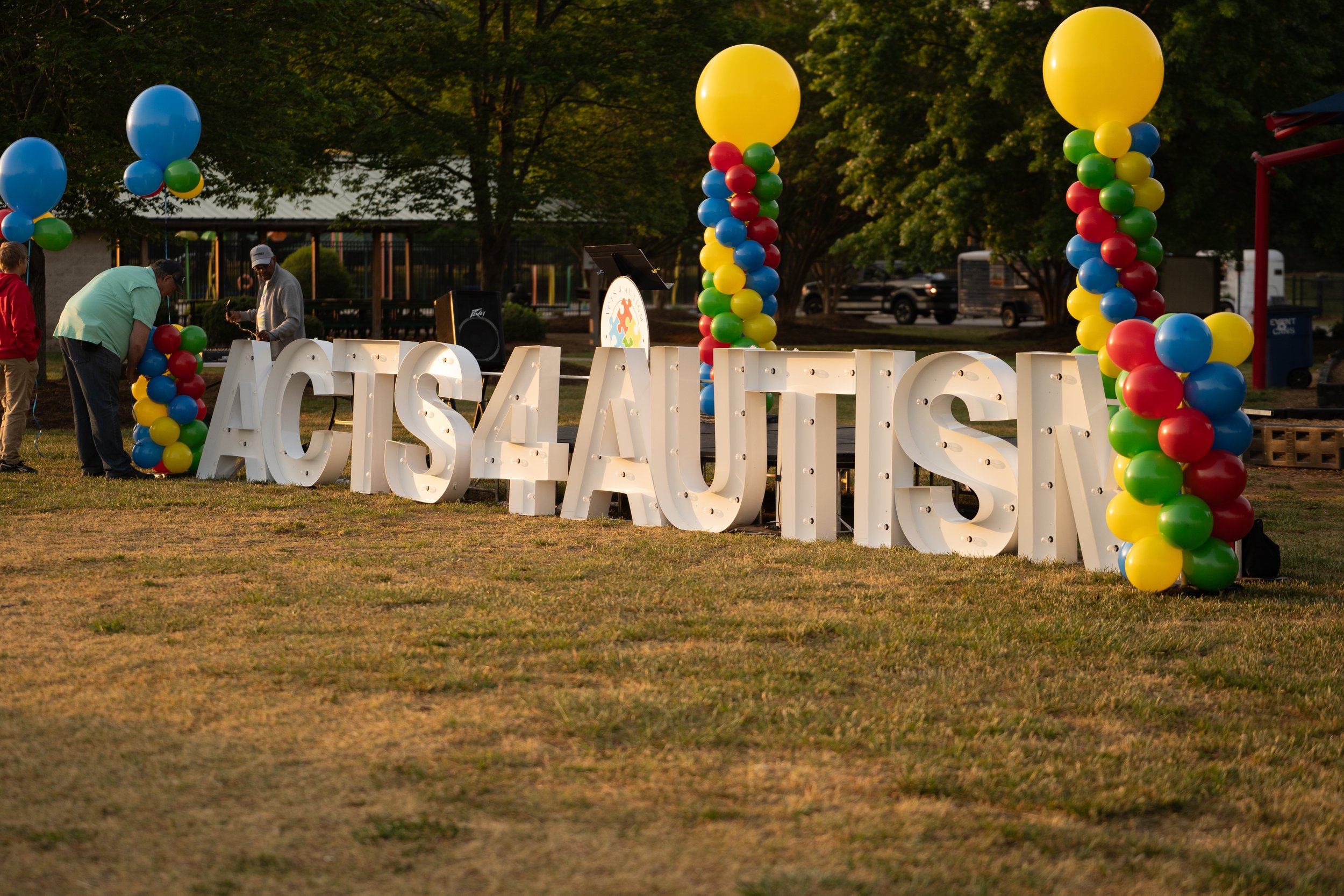 Decorative outdoor event setup with large white letters spelling 'ECS L.A.' decorated with colorful balloon columns in yellow, red, blue, and green. In the background, there are trees and people preparing for the event.