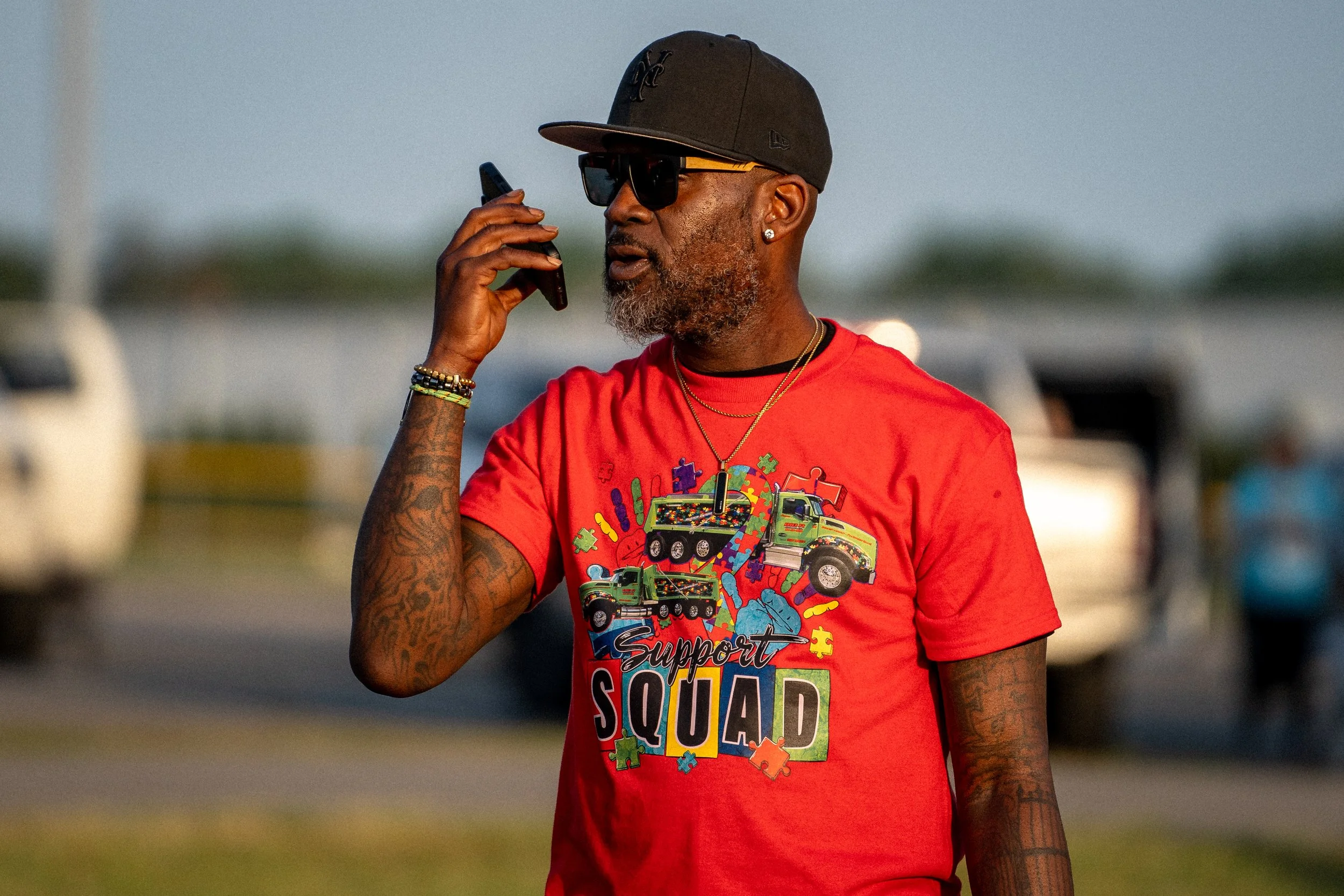 A man with tattoos and a beard, wearing sunglasses, a black baseball cap, and a red t-shirt with a colorful graphic and the text 'Support Squad', is holding a phone to his ear and standing outdoors.