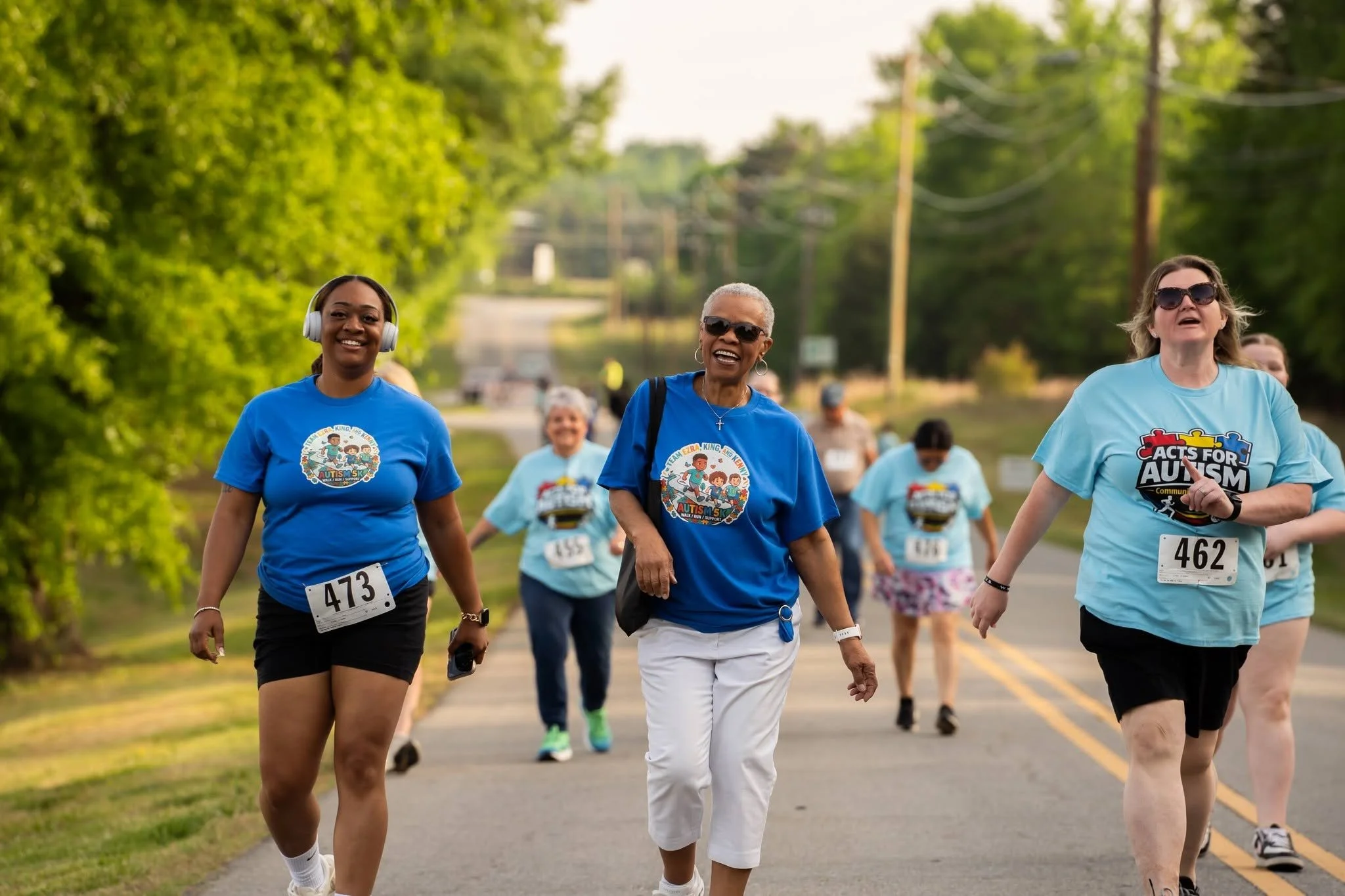 Group of women participating in a walkathon for autism awareness, walking on a suburban street surrounded by trees, some wearing blue shirts, and smiling.