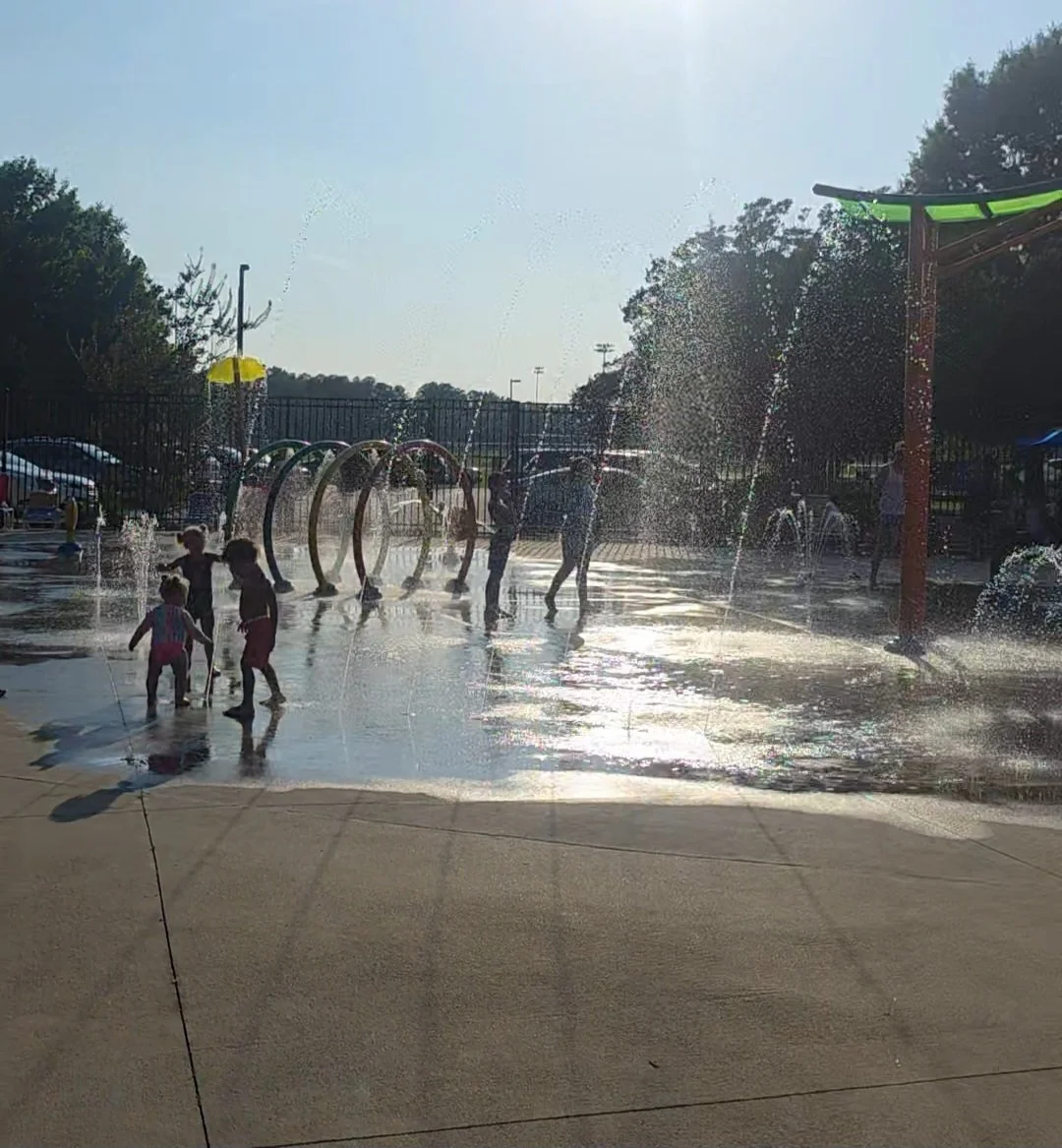 Children playing in a splash pad with water spray jets and hula hoops during a sunny day.