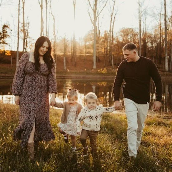 A family of five walking outdoors near a pond with autumn trees in the background, holding hands and smiling at sunset.