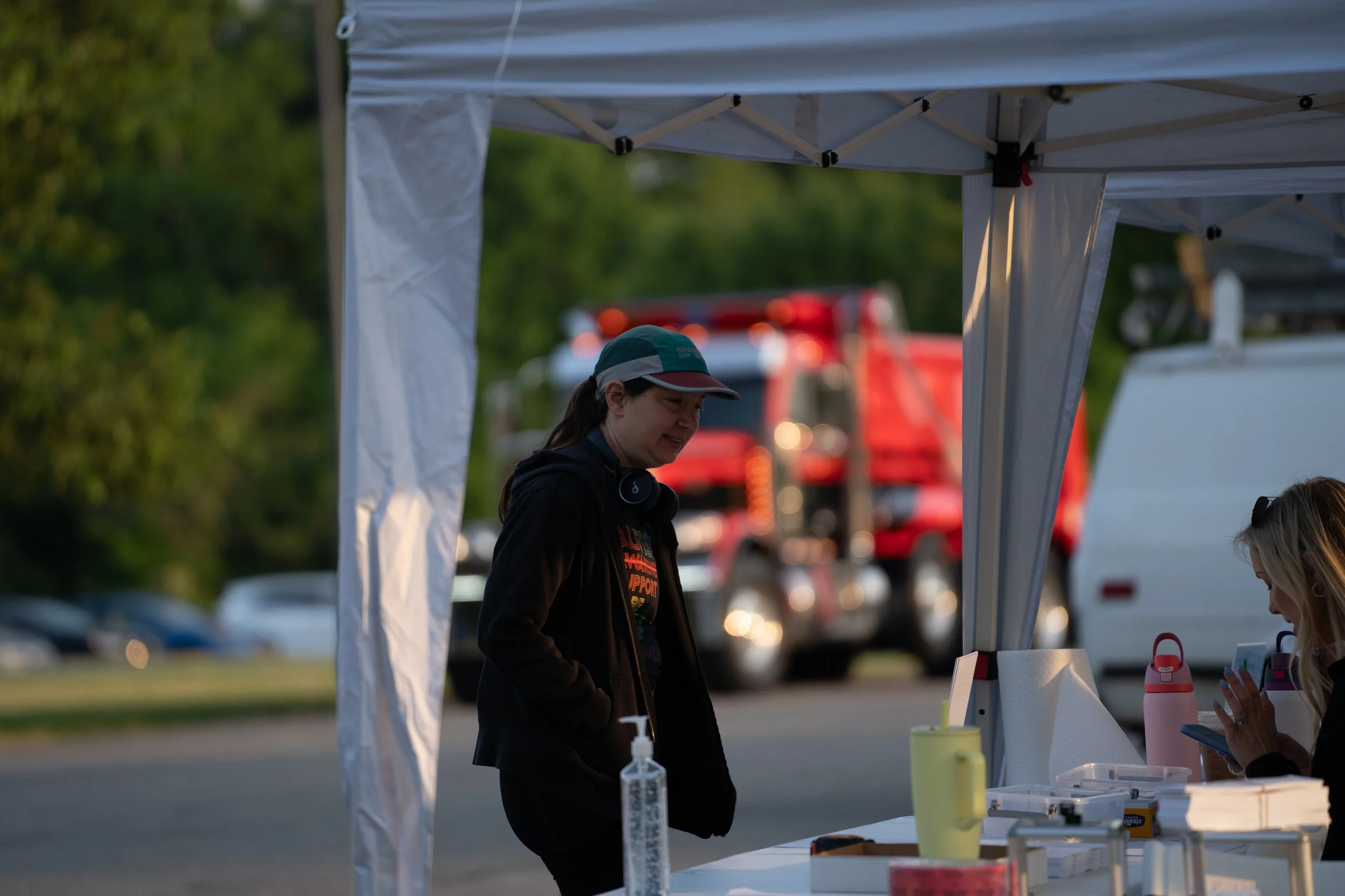 A woman in a green cap and black jacket standing under a white tent at an outdoor event. The tent is supported by metal poles, with a red truck and greenery visible in the background. There are bottles and containers on a table inside the tent, and a