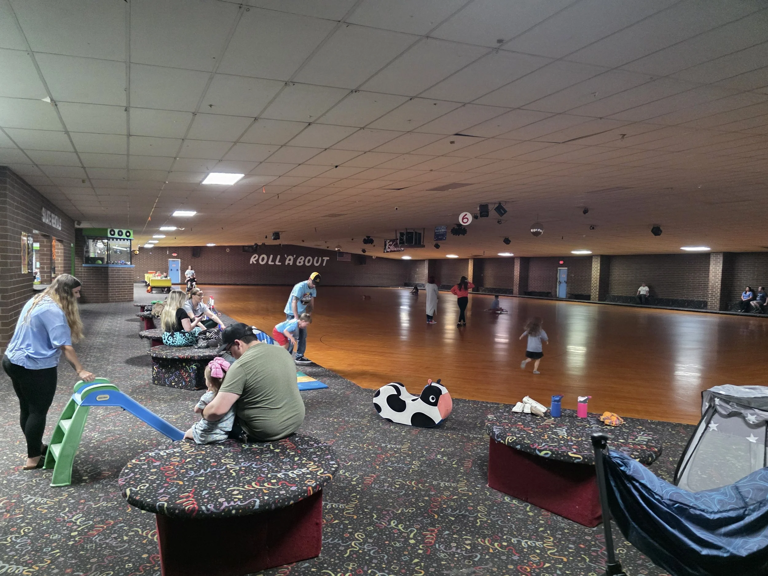Indoor skating rink with people sitting and skating, decorated with colorful confetti-themed carpet, a small slide, and a cow-shaped decoration.