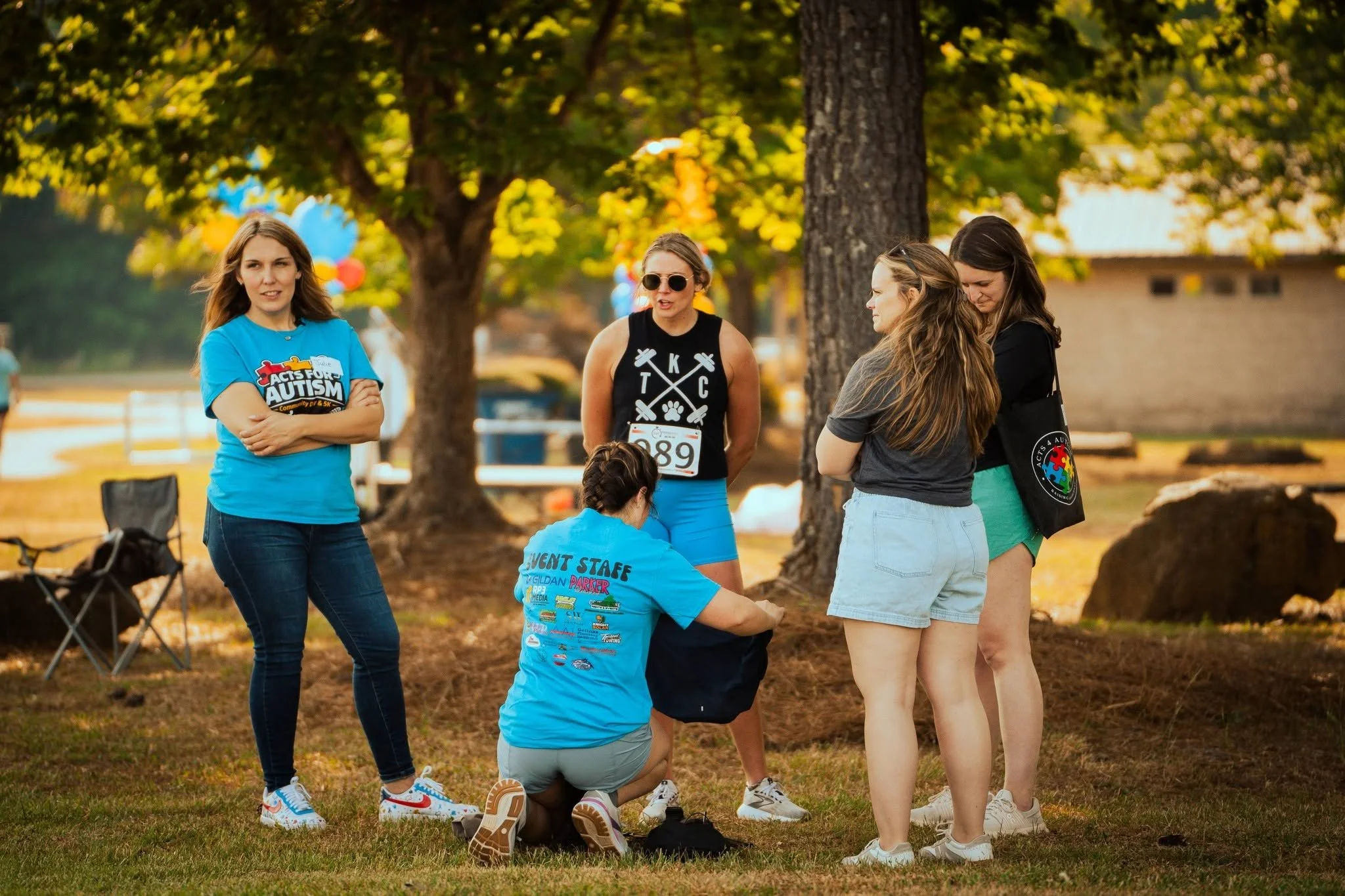Group of women gathered outdoors under trees during a community event, with some wearing event T-shirts and numbered bibs, indicating participation in a walk or run for autism awareness.